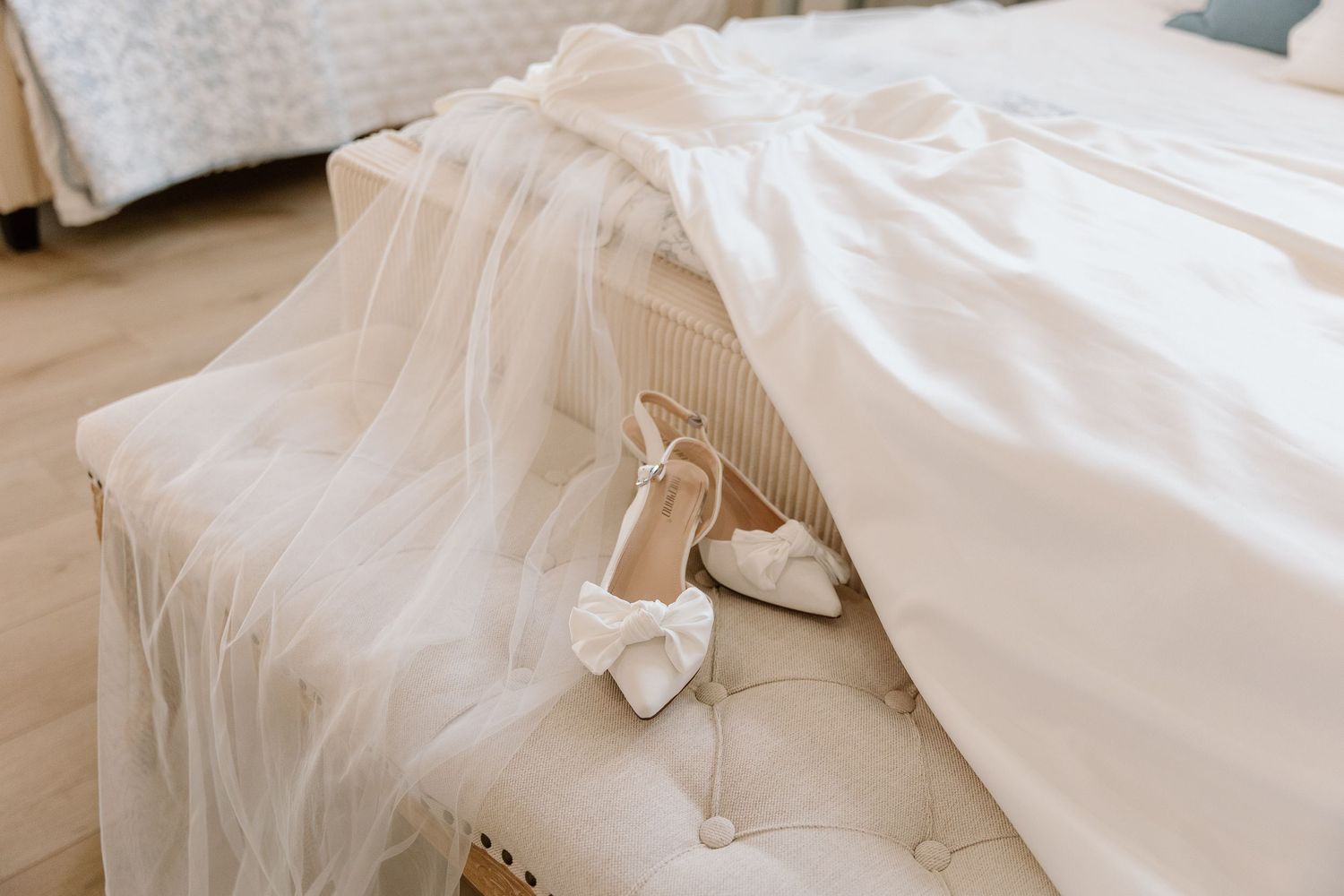 White wedding dress and bow-detail bridal heels arranged elegantly on a tufted bench beside a bed.