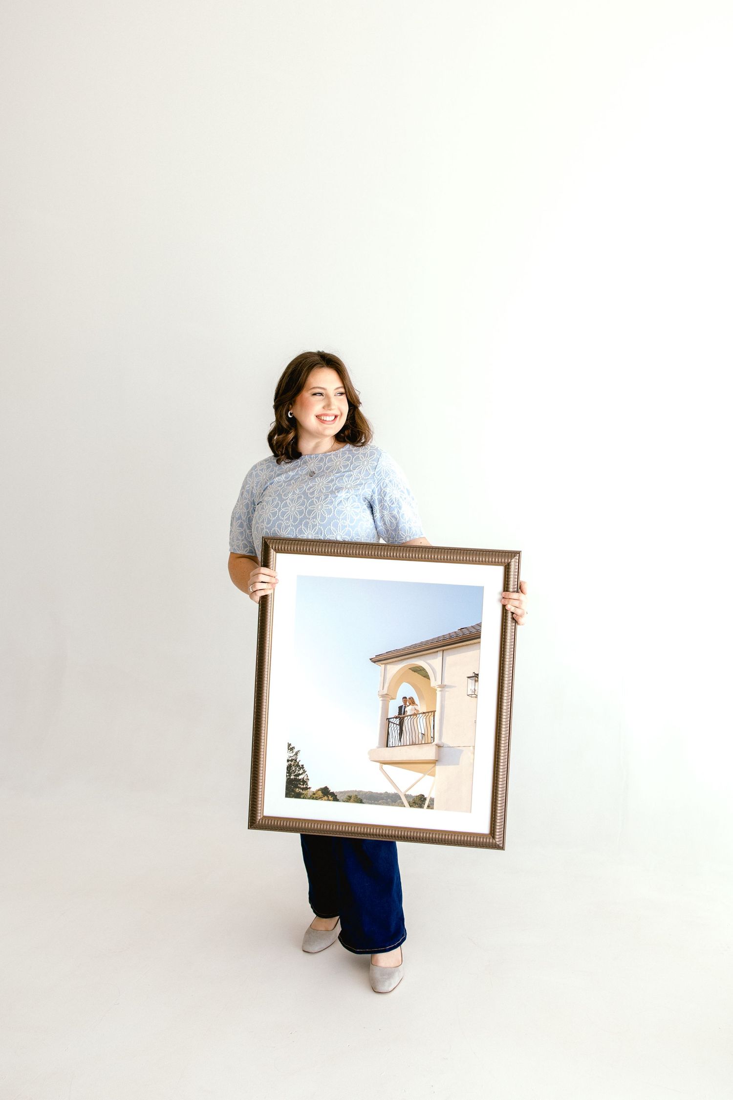 Woman smiling and holding a large framed photo of a building with blue sky, wearing a blue sweater and navy pants.