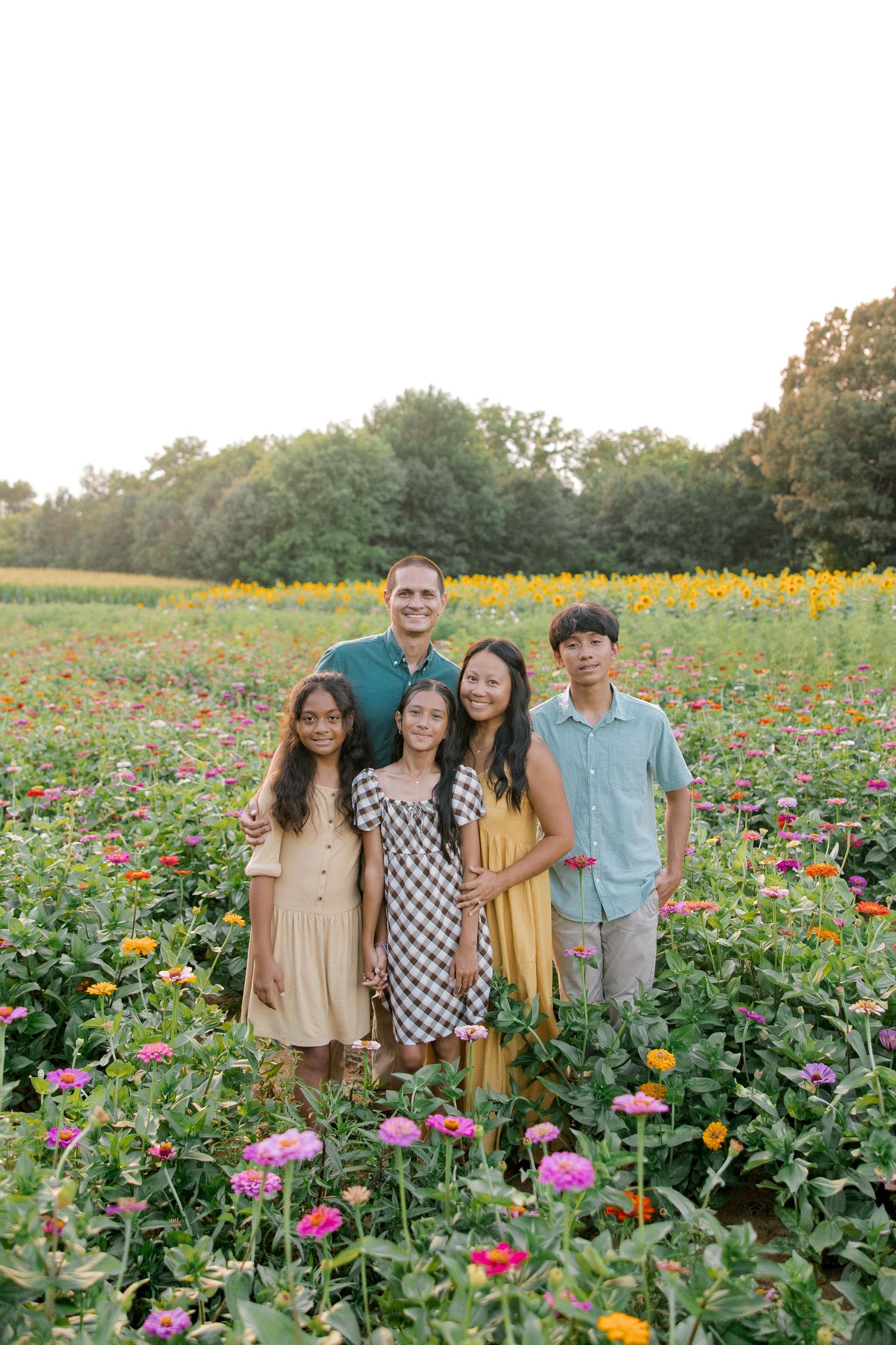 Family of five smiling together in a vibrant flower field with colorful zinnias and sunflowers in the background.