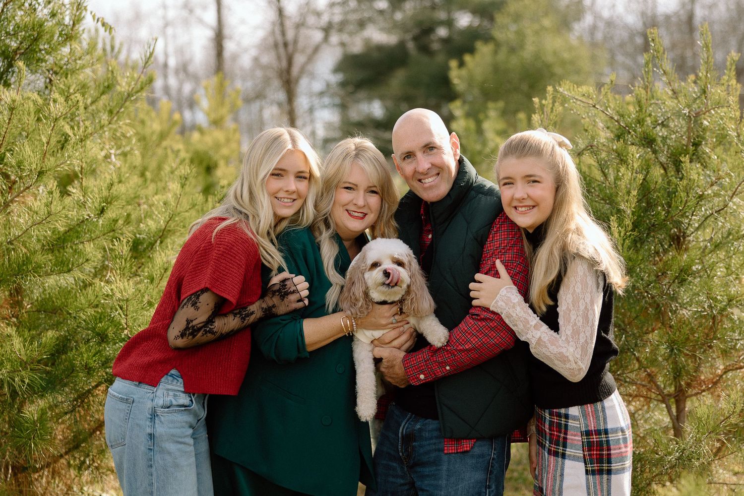 Family of four smiling with a fluffy dog among evergreen trees in a festive outdoor holiday photo session.