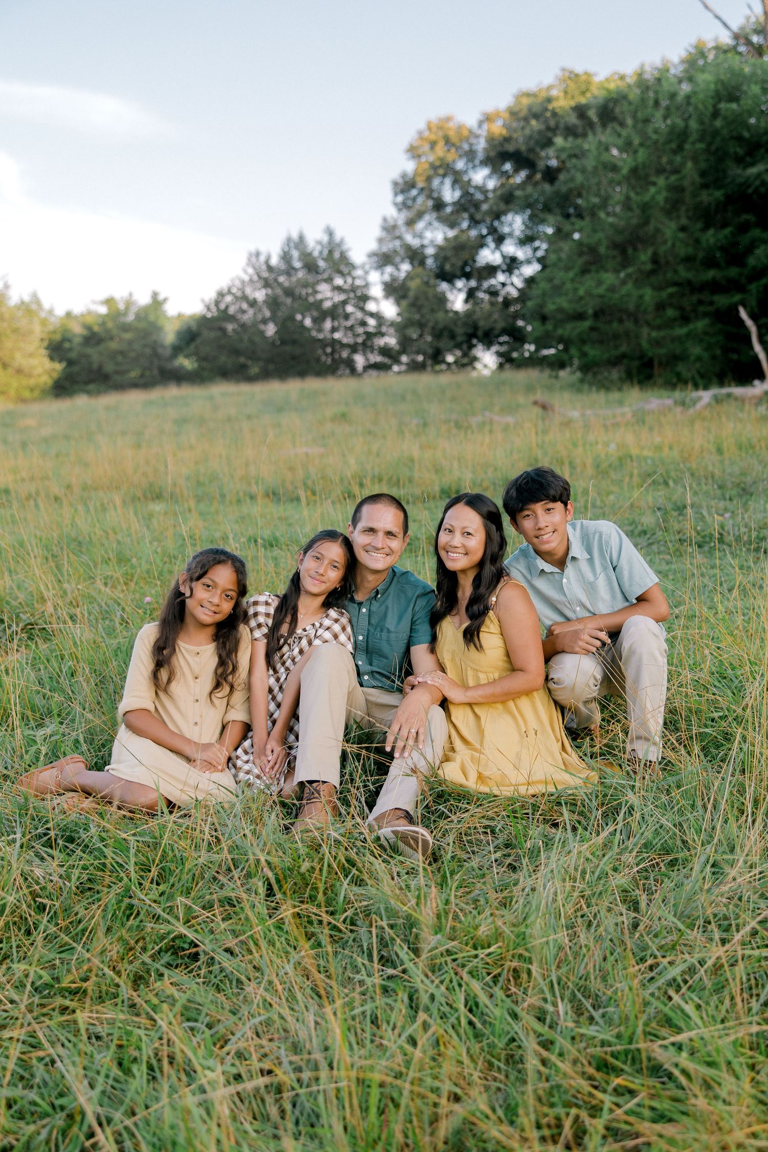 Happy family of five sitting together in a lush green meadow, smiling outdoors during golden hour.