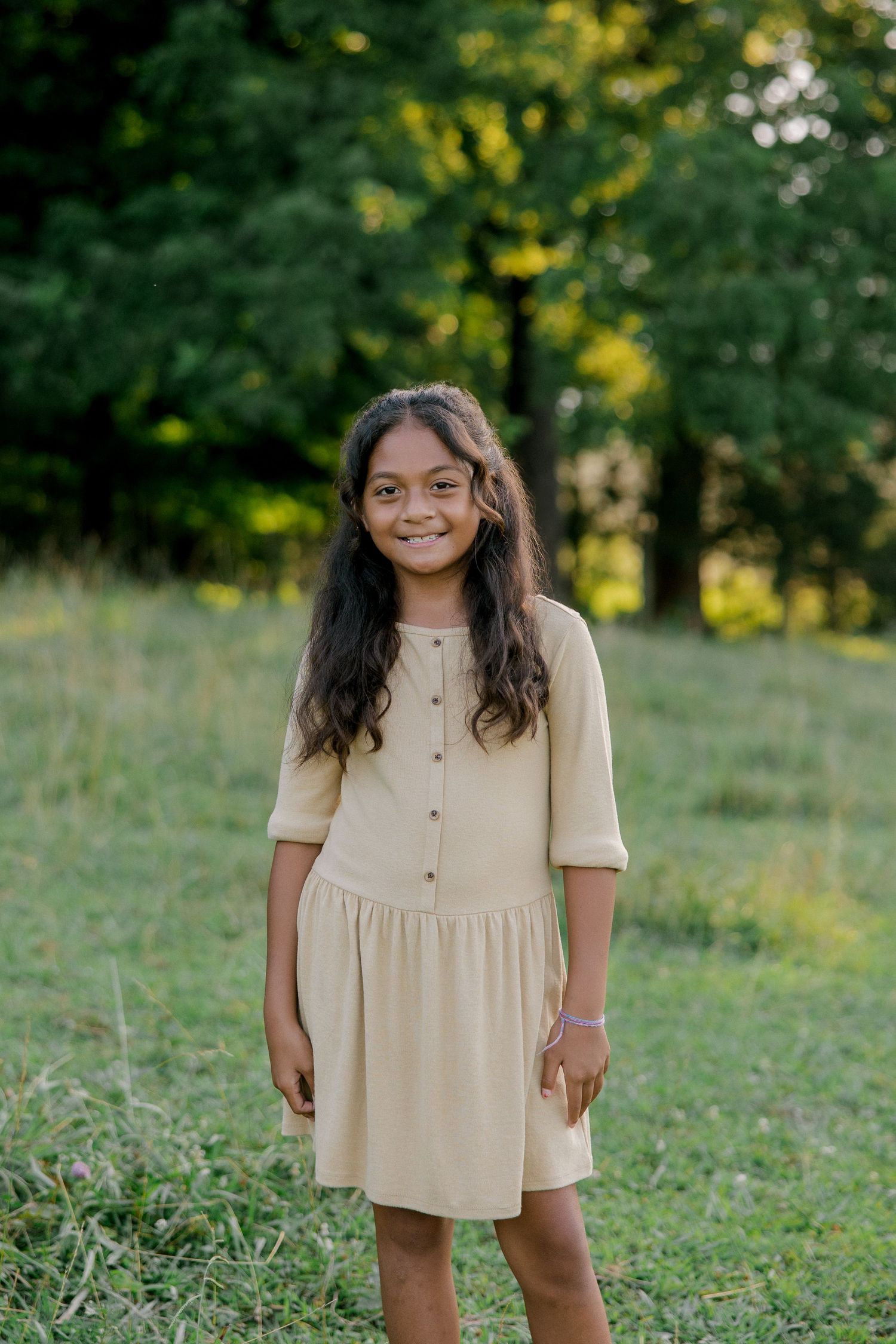 Young girl in a beige button-front dress smiling outdoors in a green grassy field with trees in background.