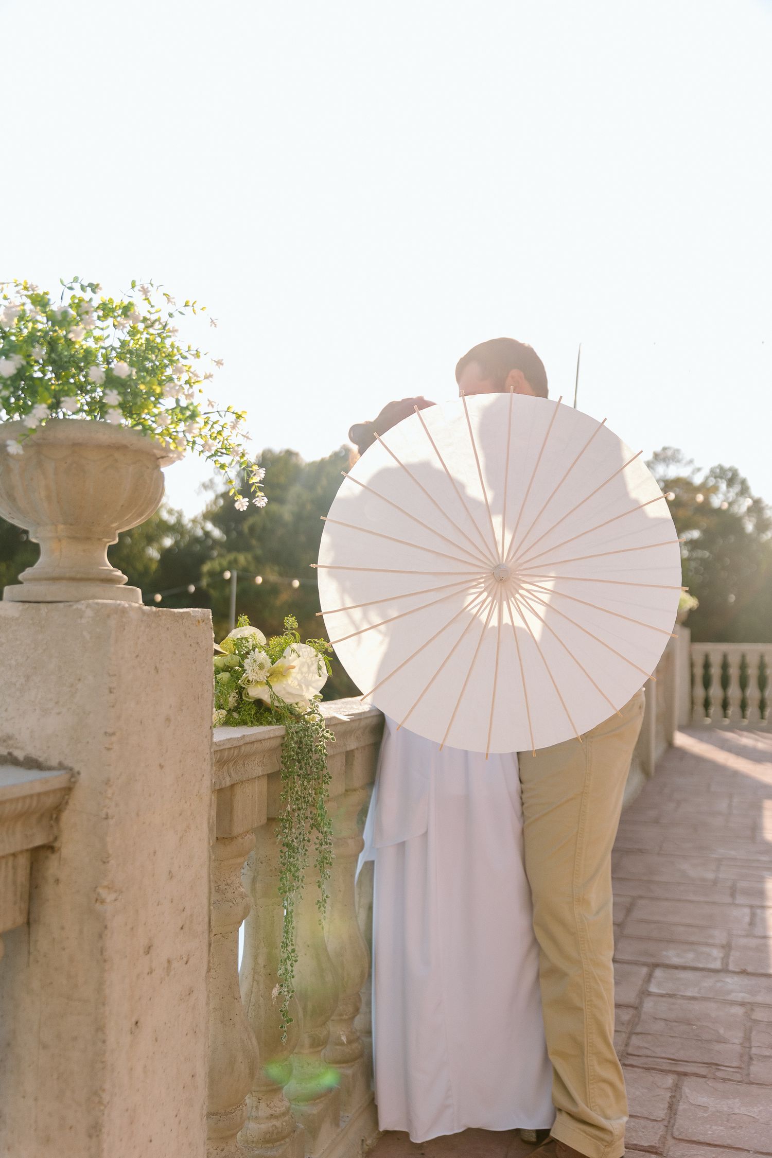 A couple shares a romantic moment on a stone terrace, partially hidden behind a white parasol, with a floral bouquet nearby.