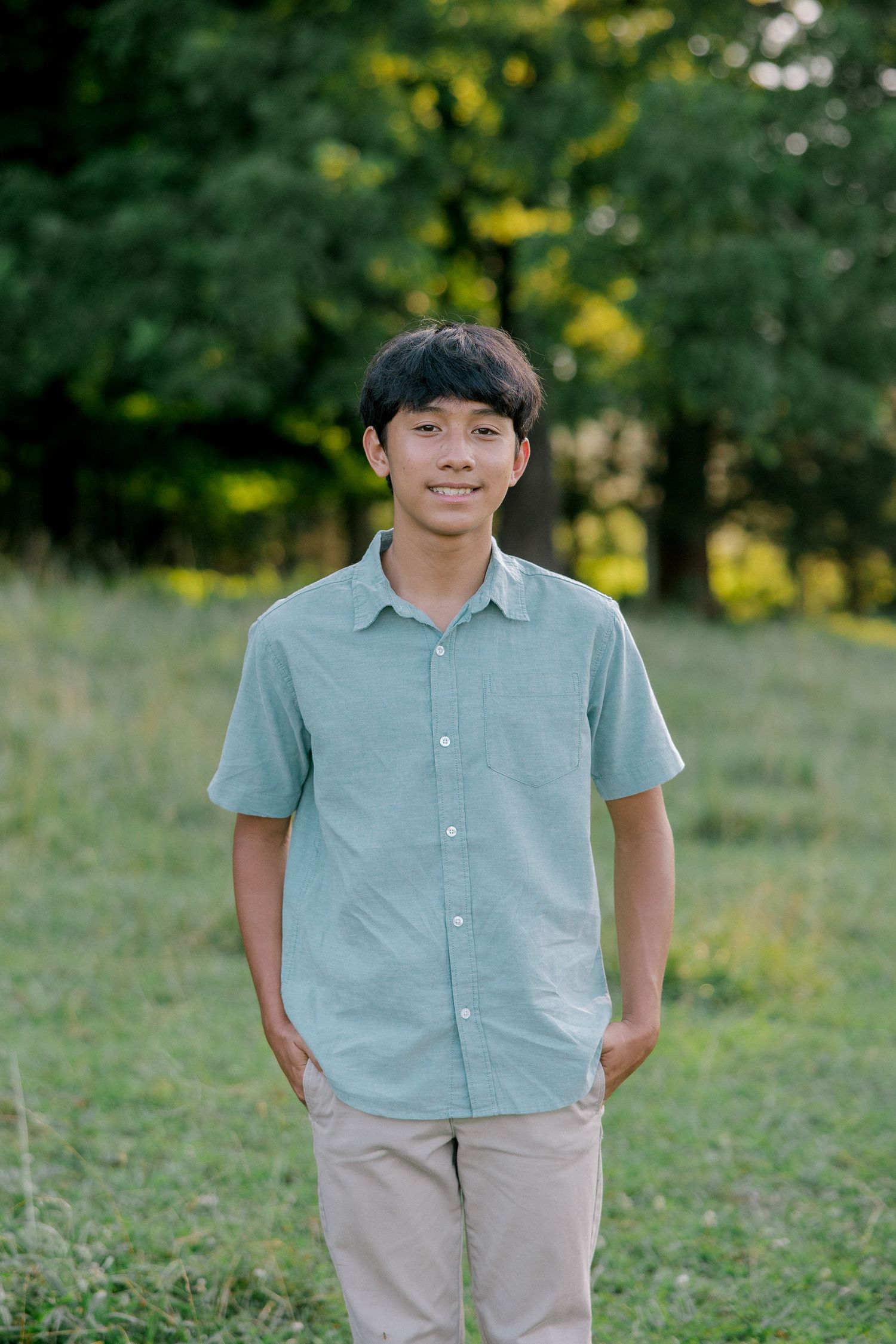 Smiling young man in a light blue button-up shirt standing in a green field with trees in the background.