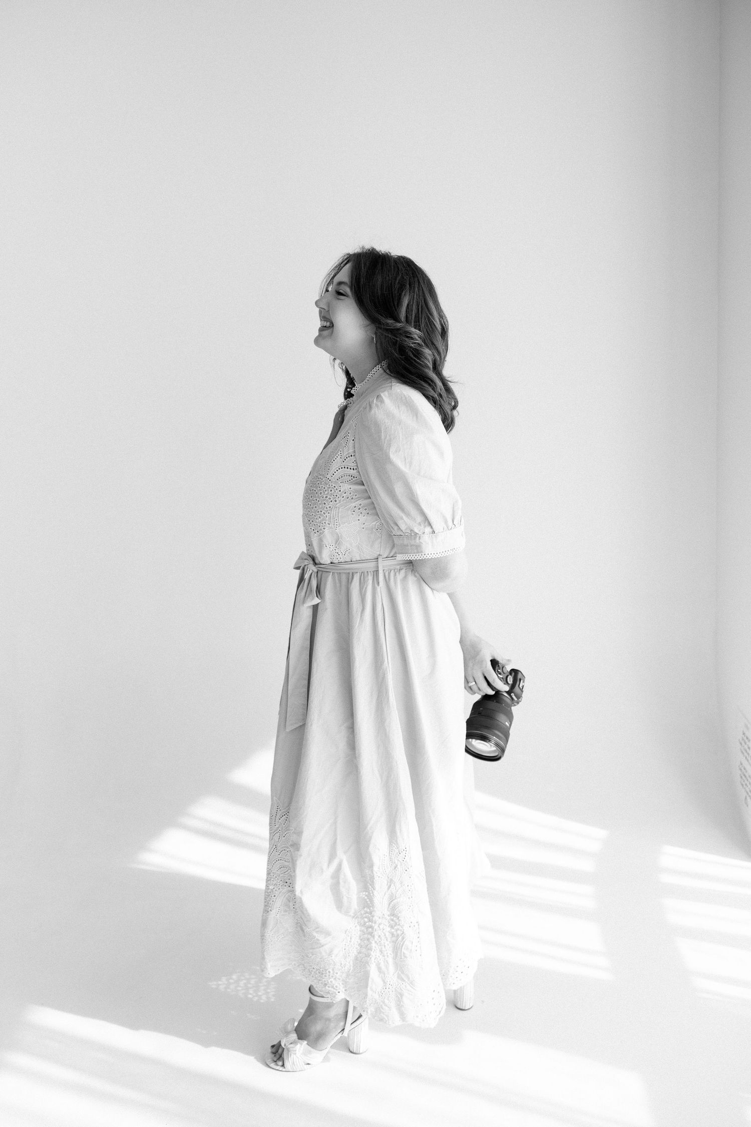 Black and white portrait of a female photographer in a flowing dress holding a camera in a sunlit studio.