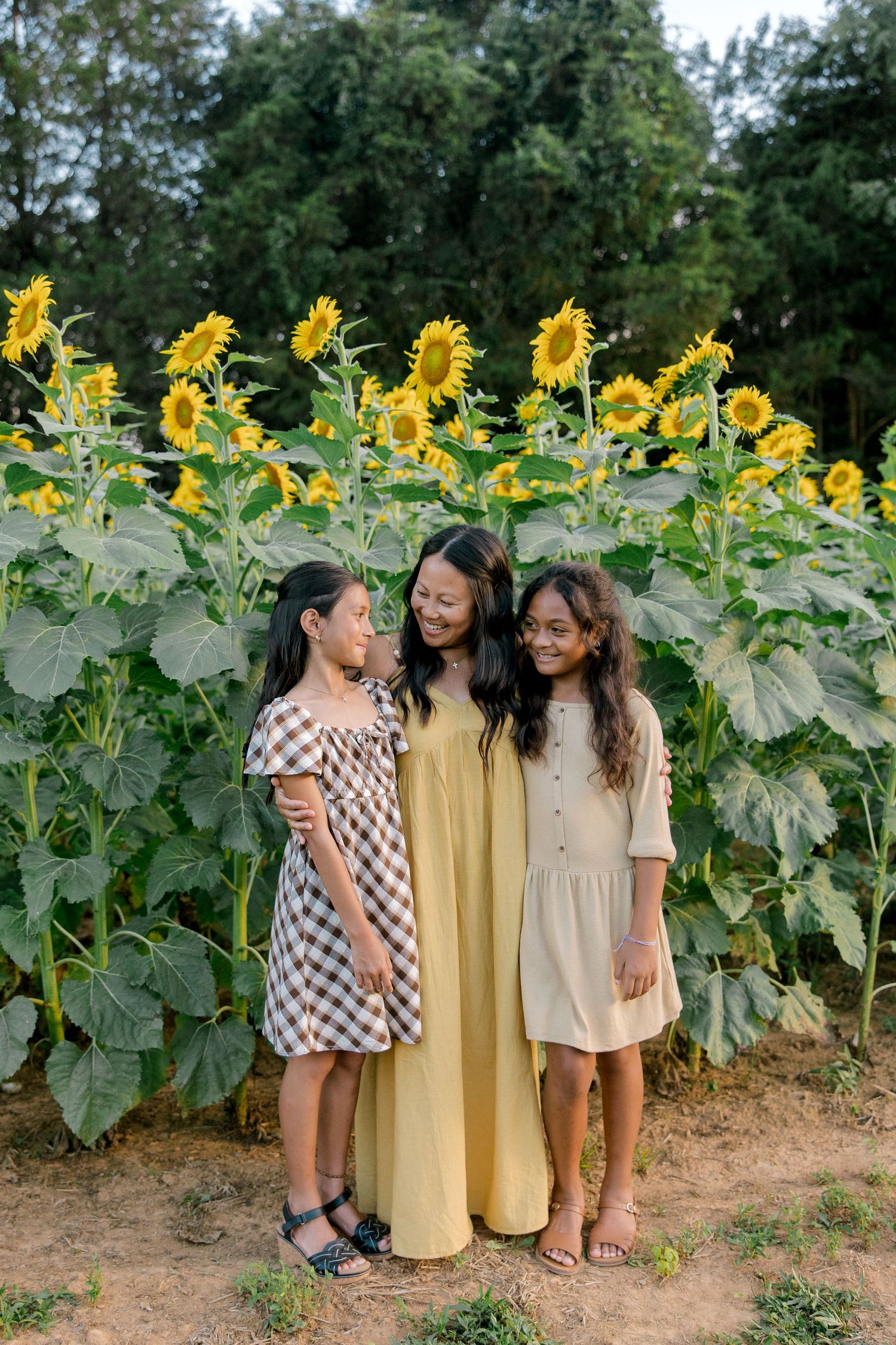A woman and two girls smile together in front of tall sunflowers in a sunny outdoor field.