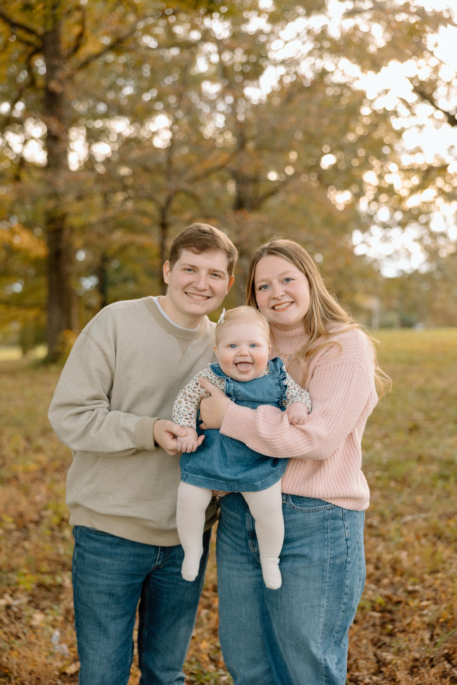 Young couple smiling outdoors in autumn park holding happy baby in denim outfit, fall foliage background.
