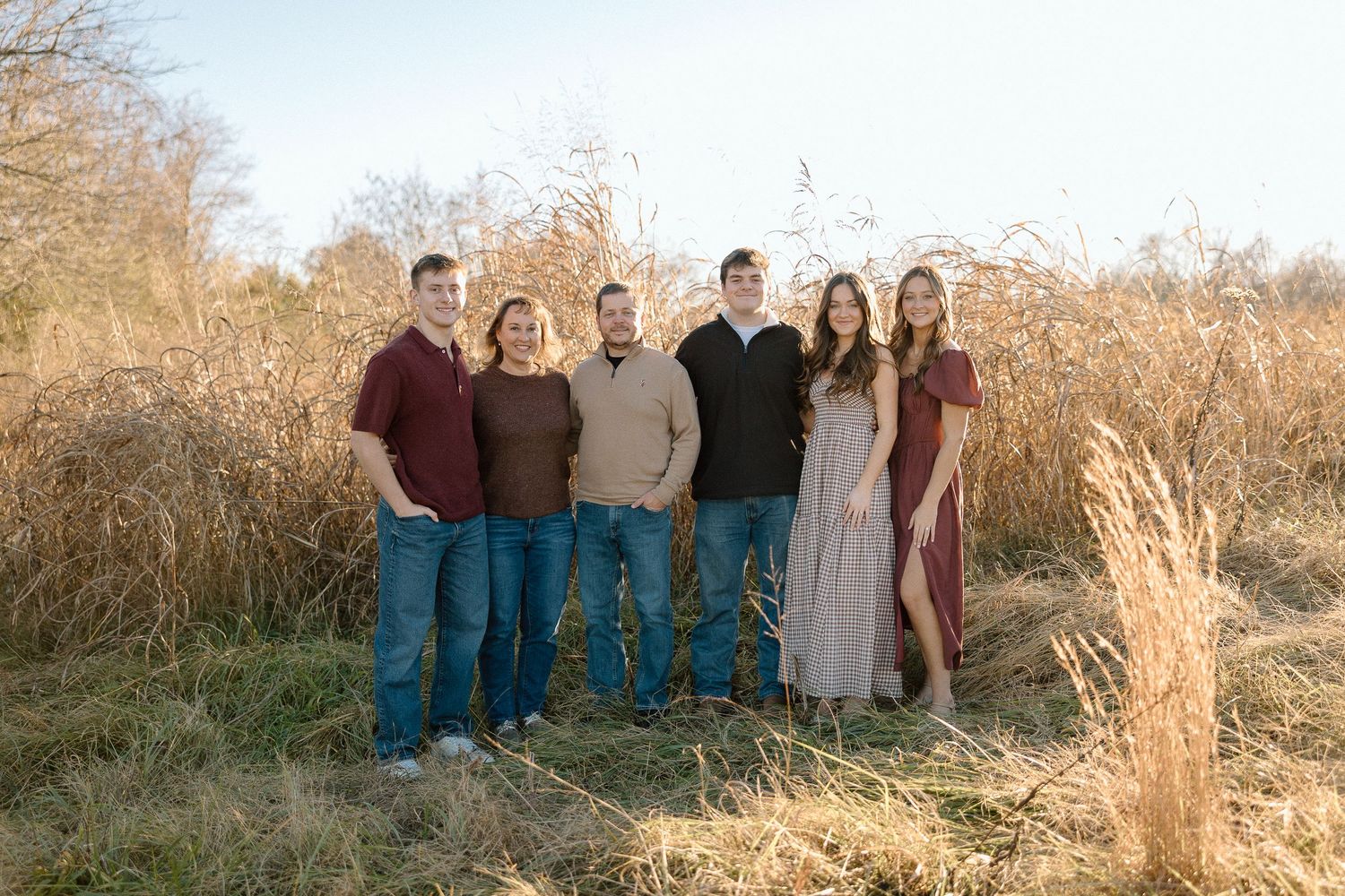 Family of six posing outdoors in fall, standing in front of tall golden grass in casual coordinated outfits.
