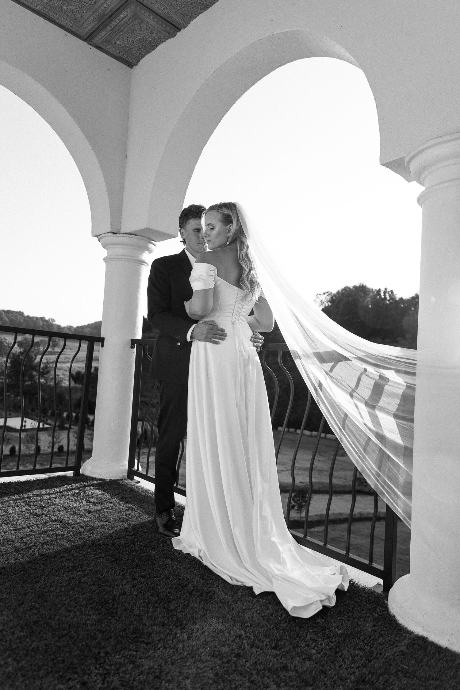 Black and white wedding photo of bride and groom embracing under elegant arched columns, veil flowing in the breeze.