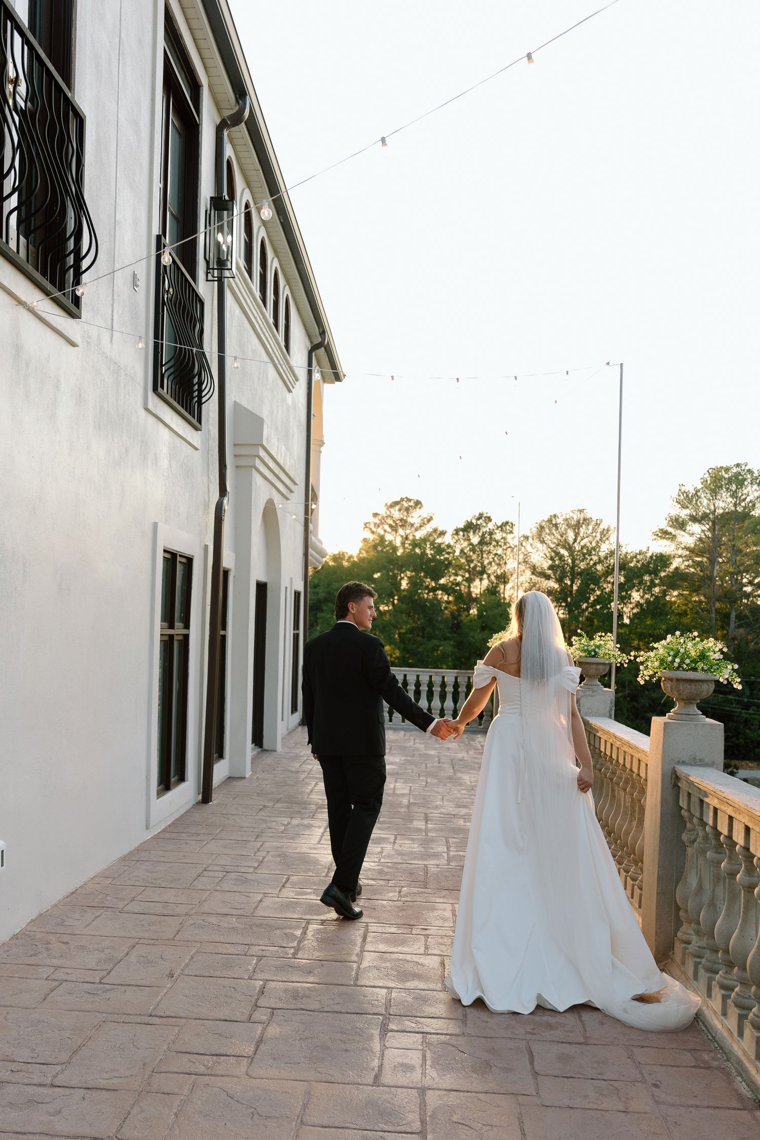 Bride and groom holding hands walking along elegant outdoor terrace at golden hour, bride in white gown with veil.