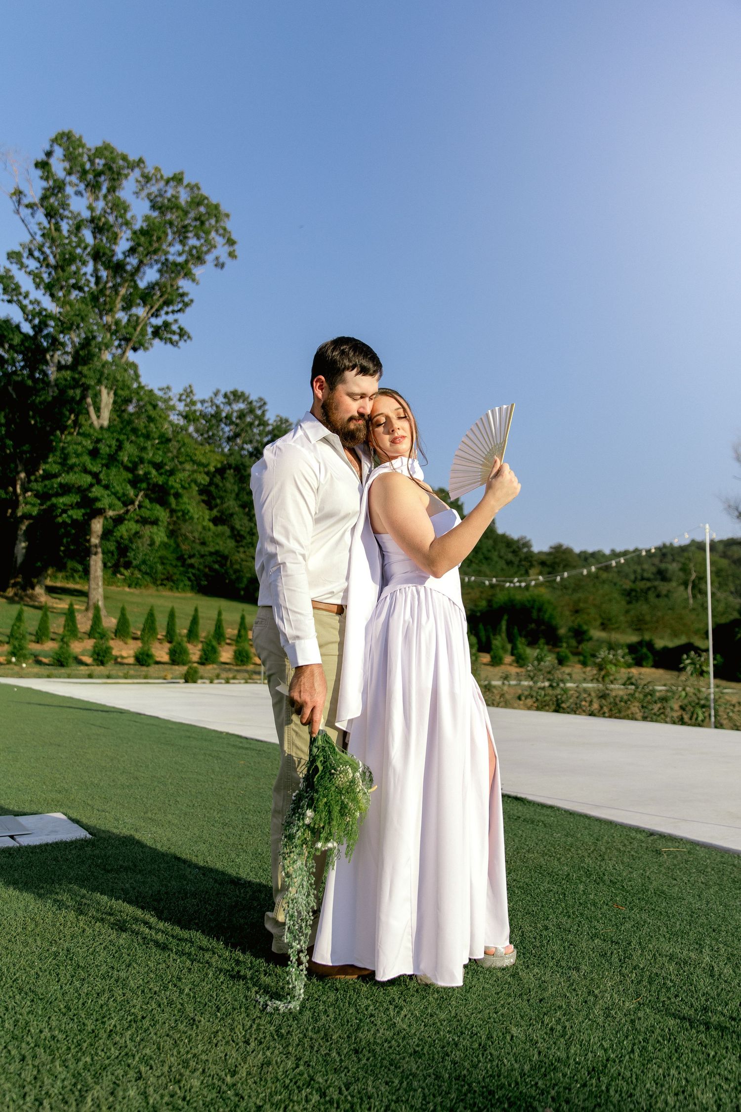 A couple in white attire embraces outdoors on green grass, bride holding a fan and groom holding a bouquet.