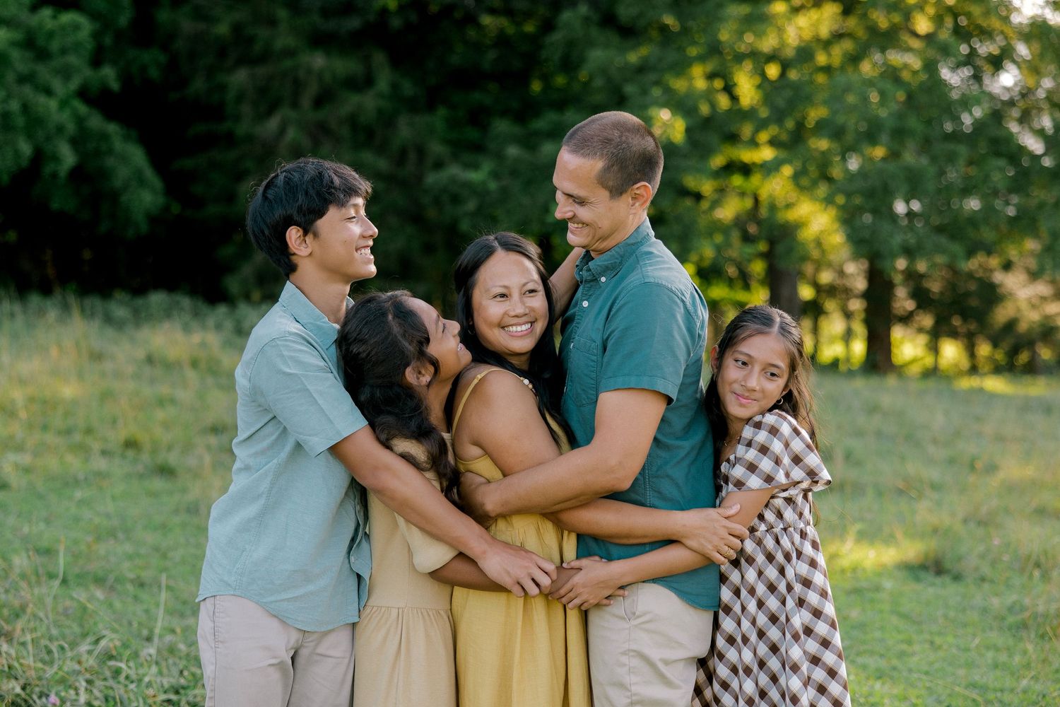 Family of five hugging and smiling together in a green field during golden hour outdoor photo session.