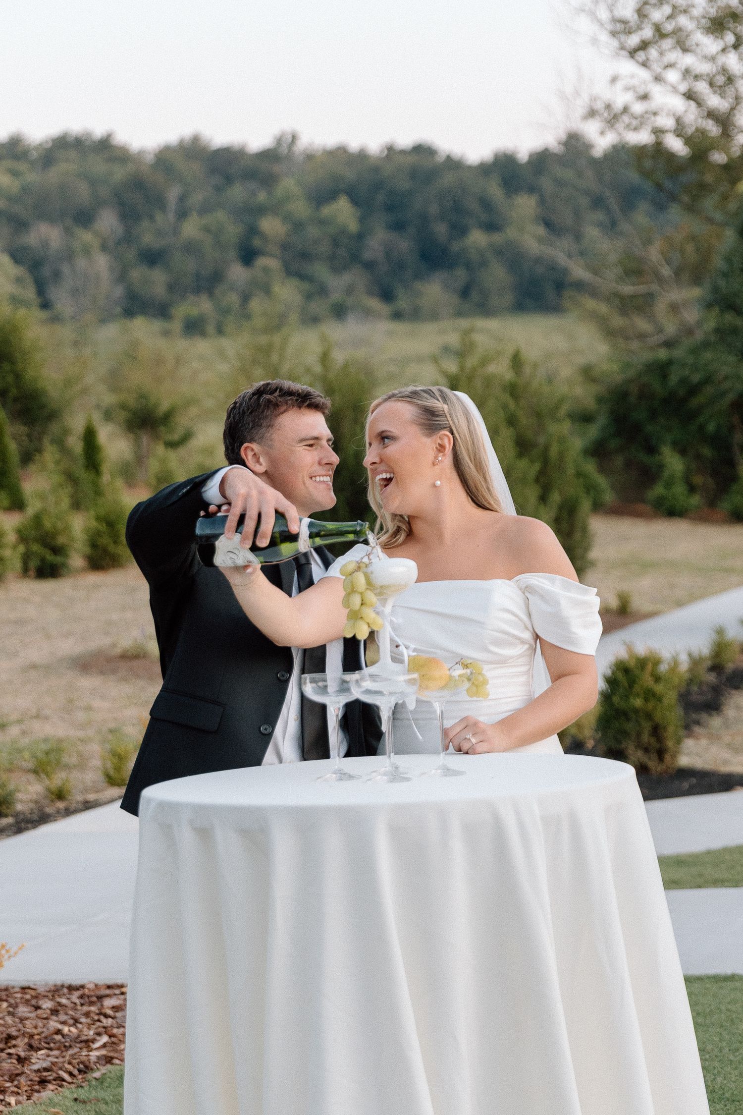 Bride and groom pouring champagne tower at outdoor wedding reception with lush green landscape backdrop.