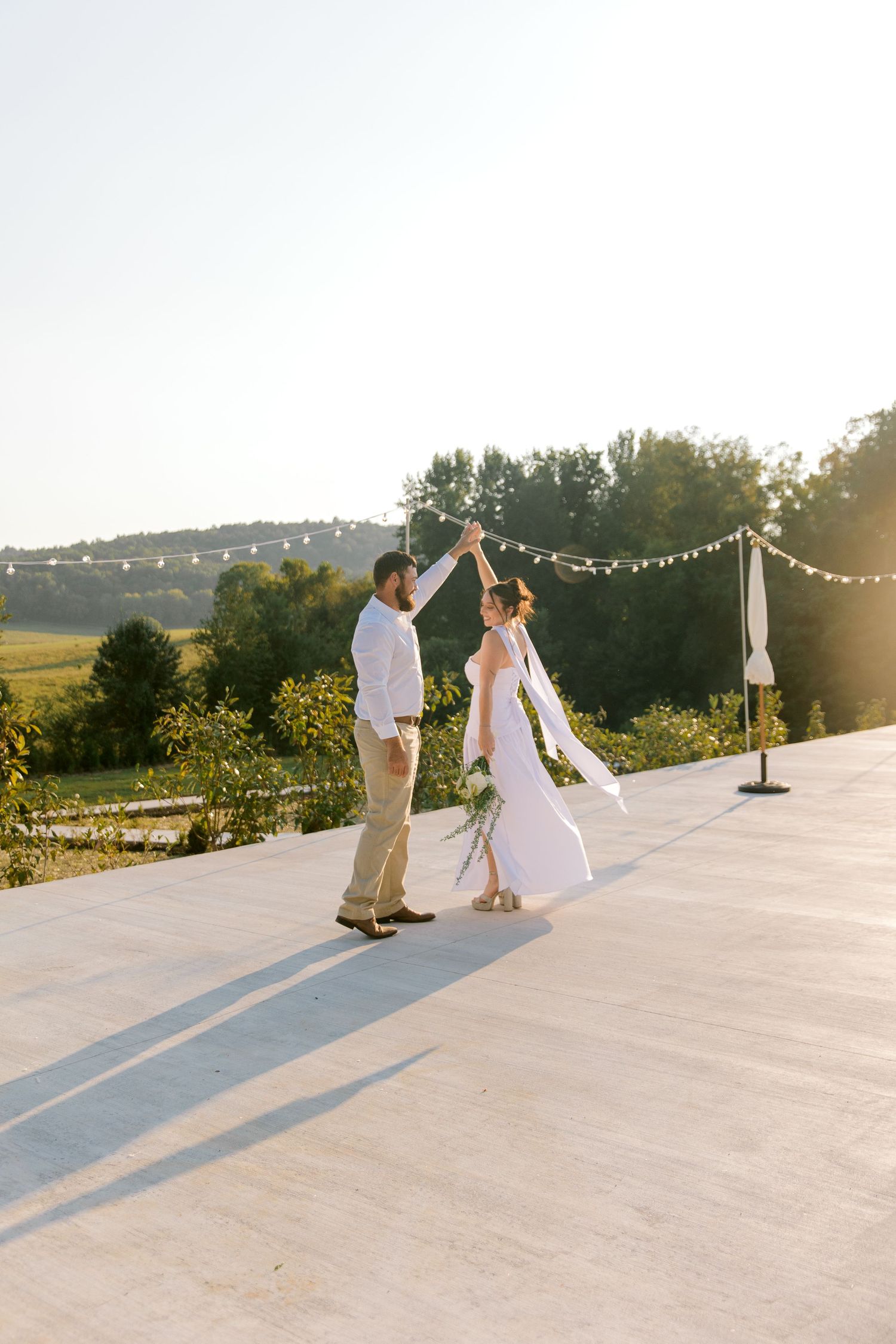Bride and groom dancing outdoors at golden hour on a rooftop venue with string lights and scenic greenery.