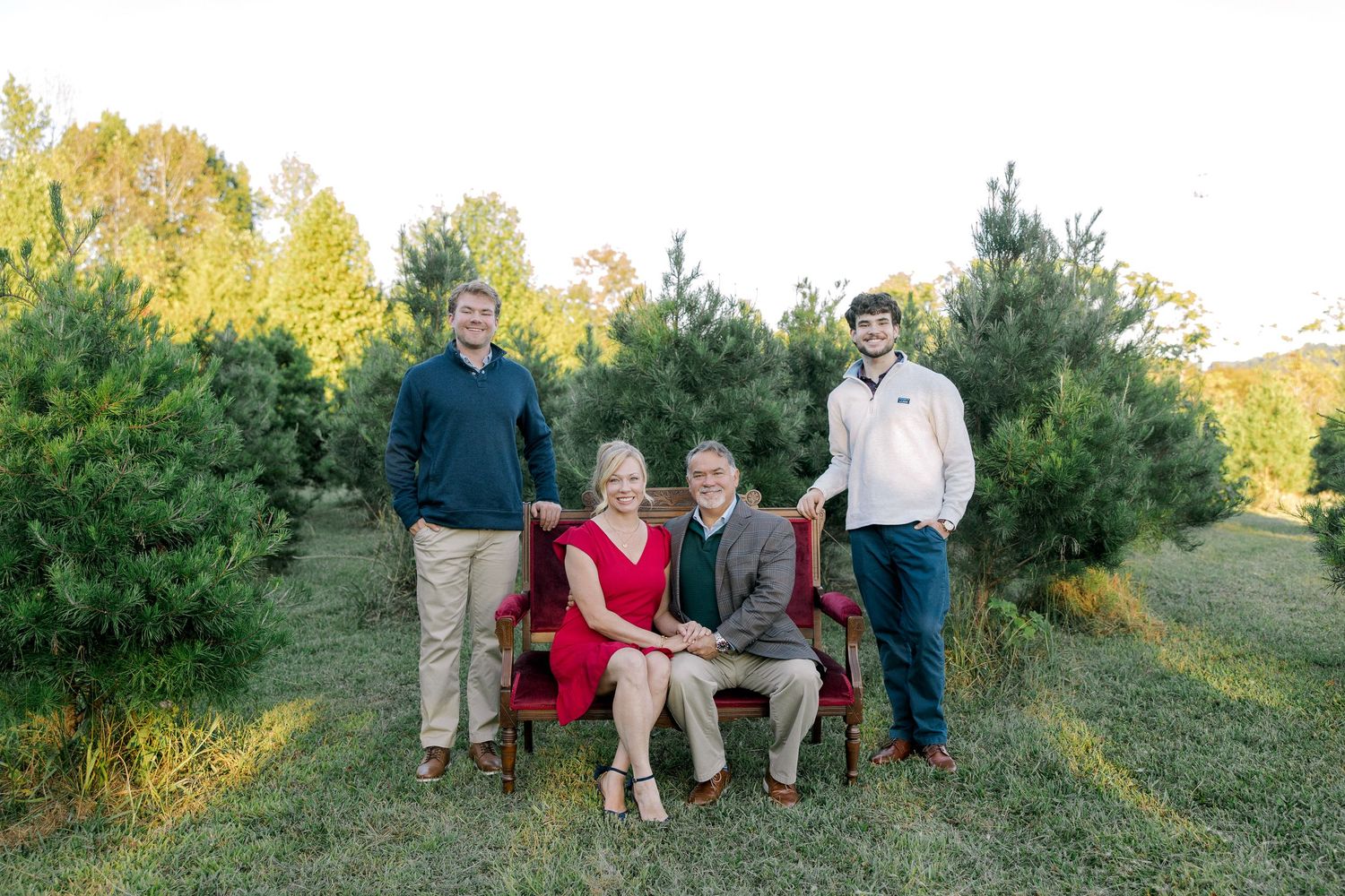 Family of four posing outdoors at a Christmas tree farm, two sons standing behind seated parents on a red bench.