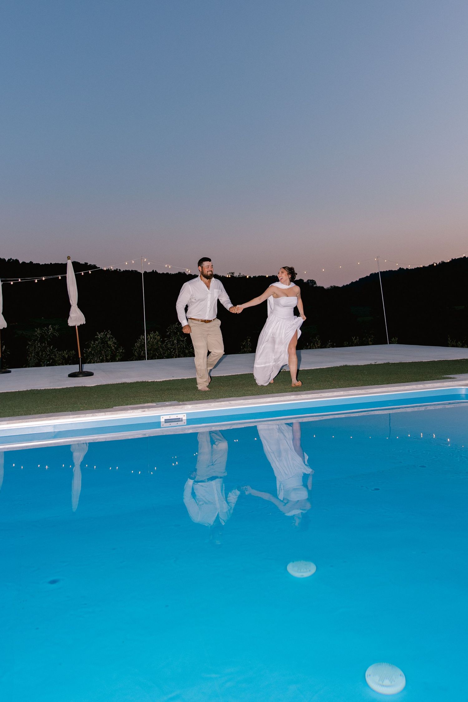 Bride and groom holding hands running by illuminated pool at dusk during outdoor wedding celebration.