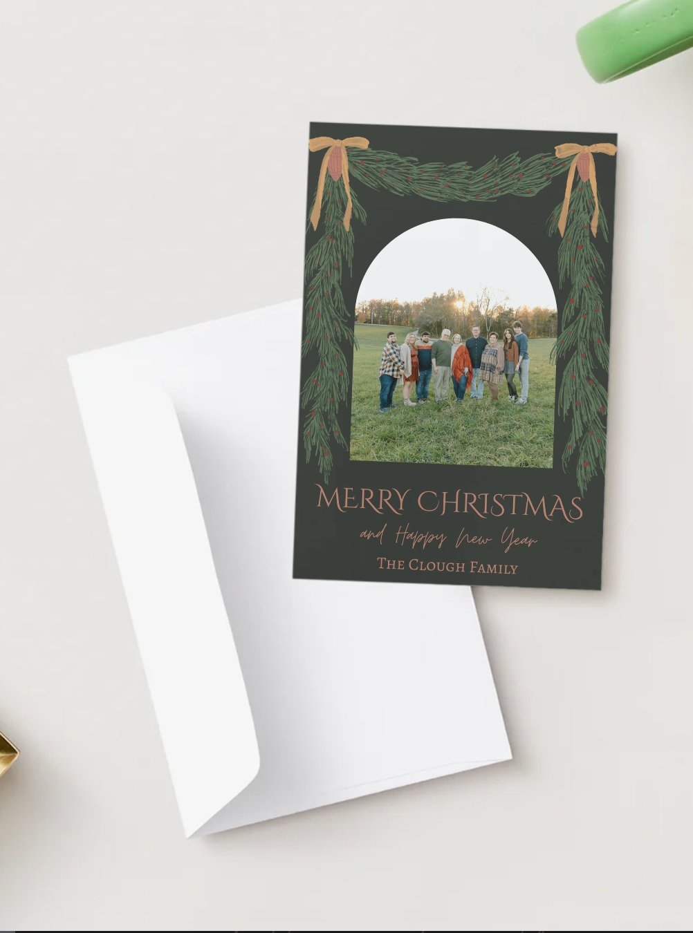 Green Christmas card with pine garland arch, family photo, Merry Christmas text, and white envelope.