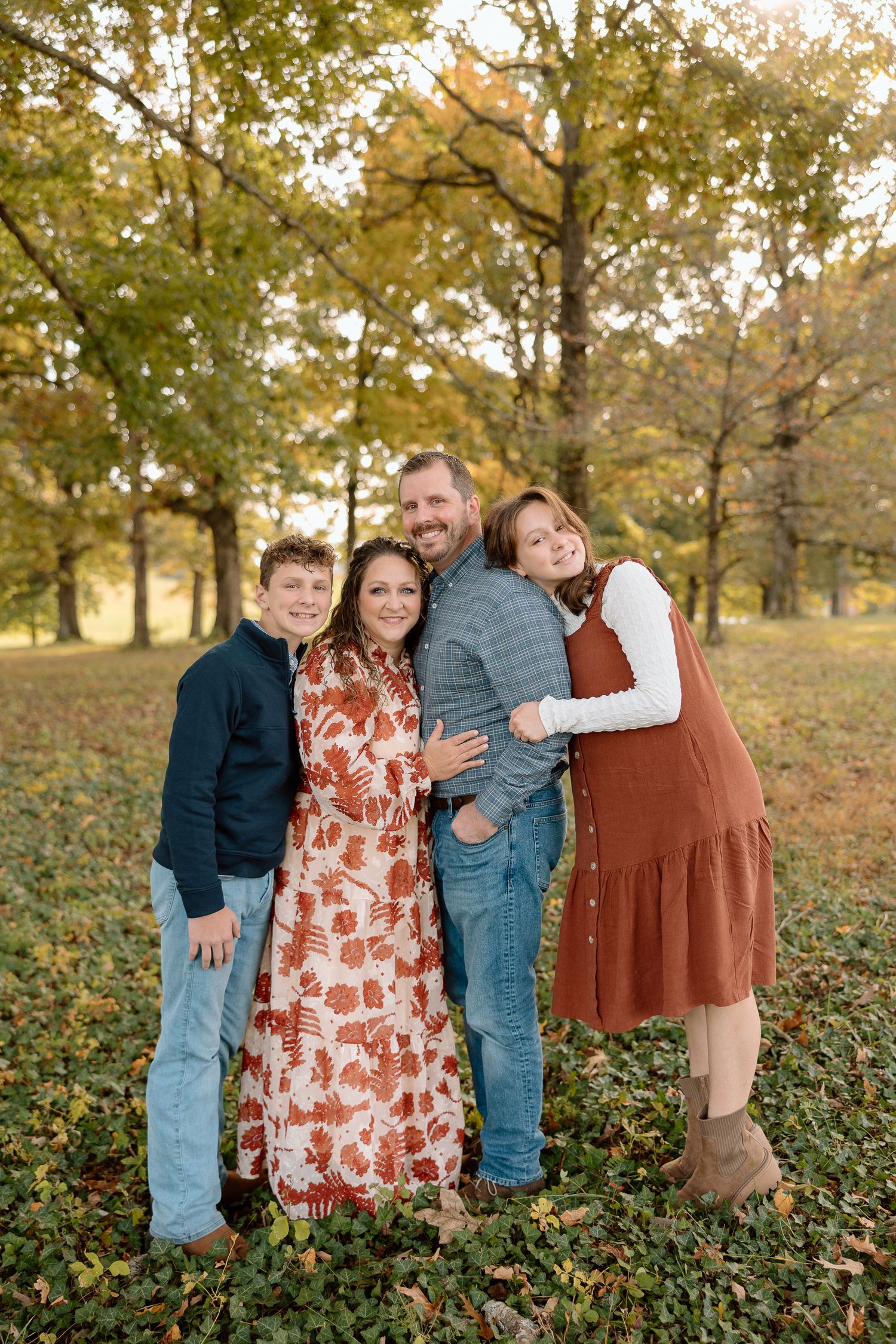 Family of four smiling together in a park during autumn, surrounded by colorful fall foliage.