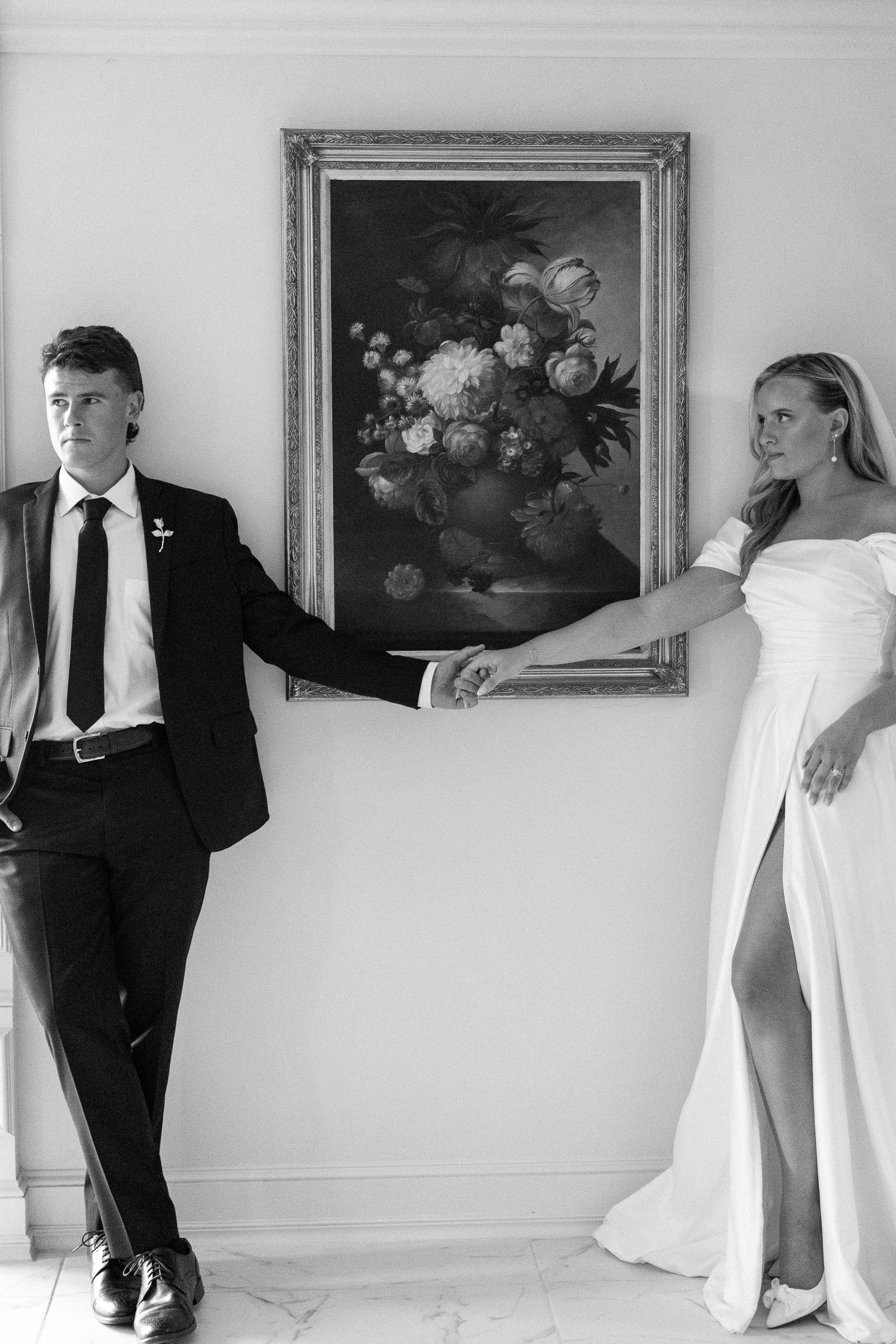 Black and white wedding photo of bride and groom holding hands beside a floral painting.