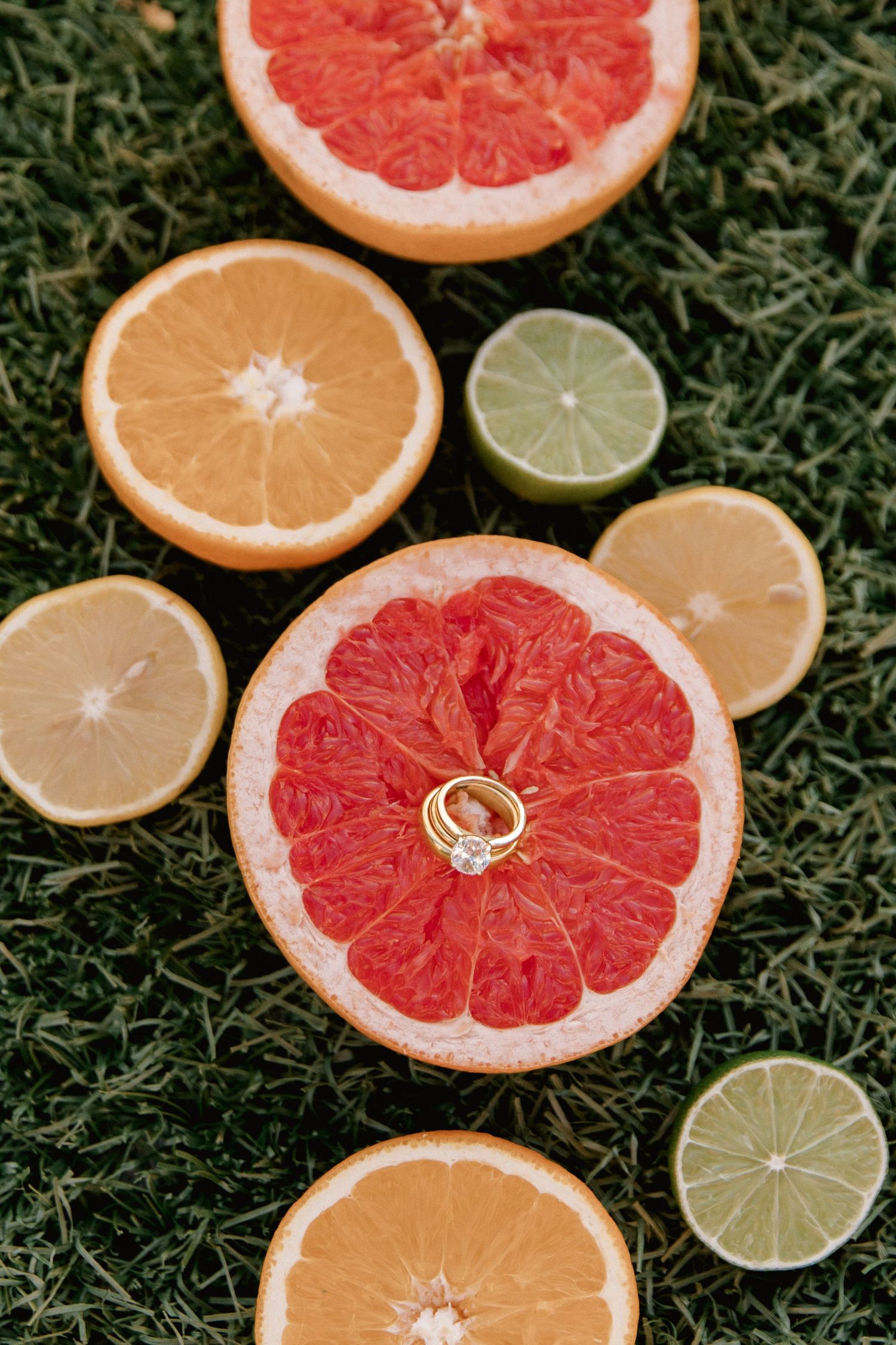 Gold engagement rings resting on a halved grapefruit surrounded by sliced citrus fruits on green grass.