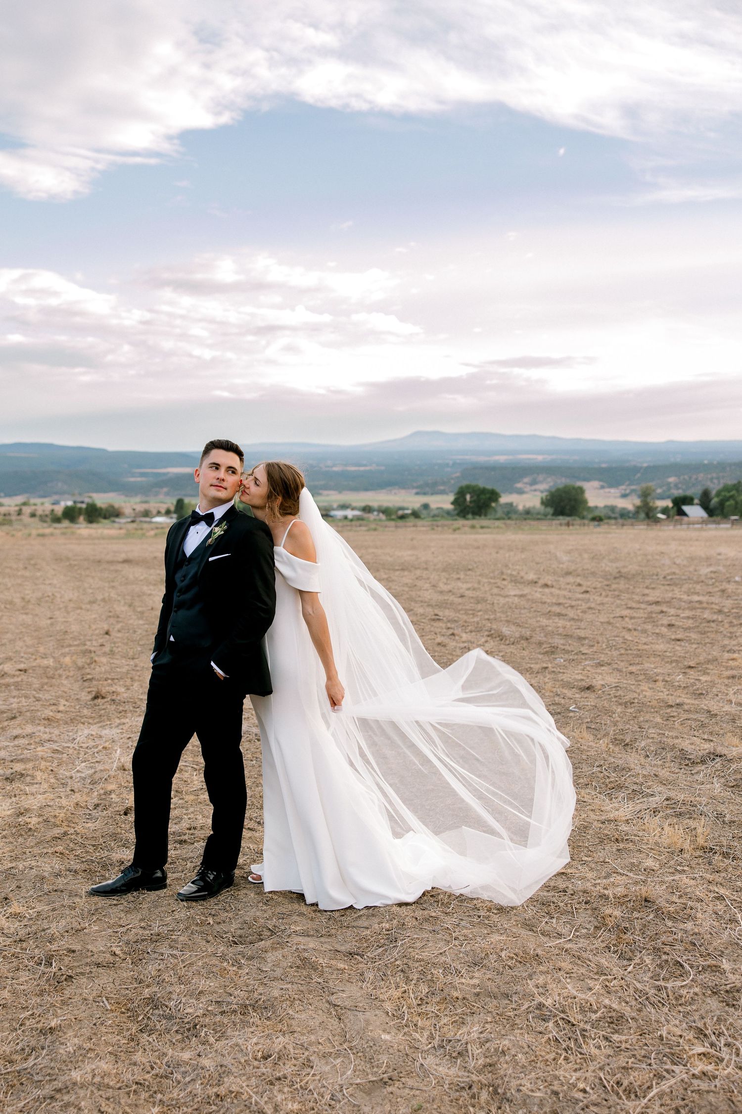 Bride kissing groom on cheek in open field with flowing veil and mountain backdrop at sunset.
