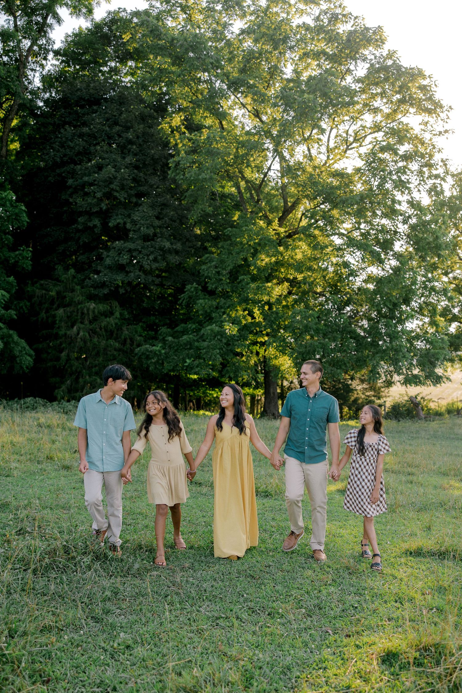 A family of five holds hands walking through a green field with trees in the background during golden hour.