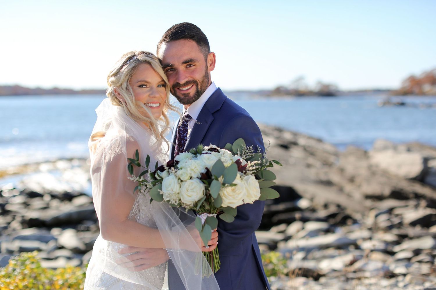Couple shares romantic moment on rocky beach with ocean and bouquet.