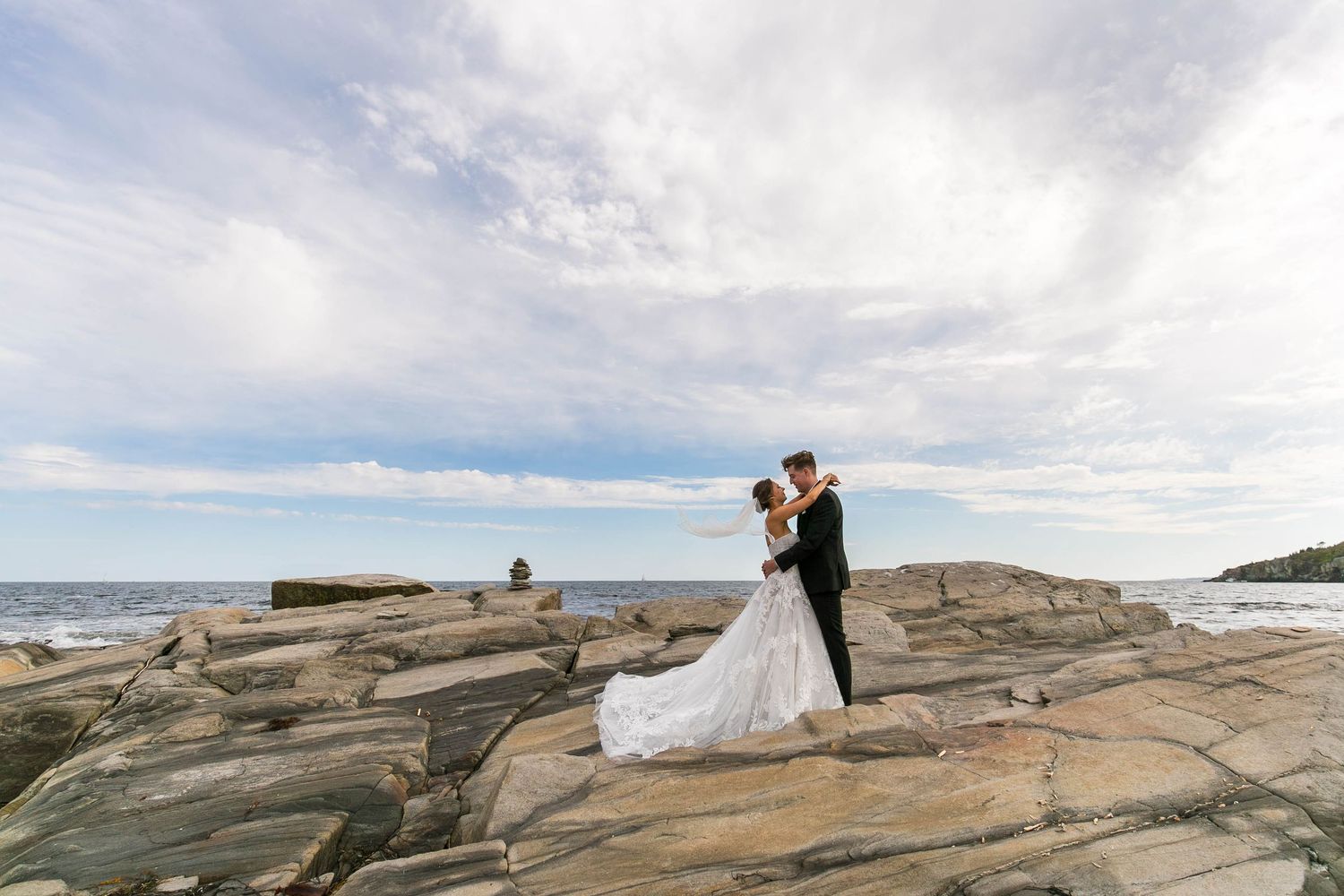 Wedding couple poses on rocky coastal shoreline under cloudy sky.