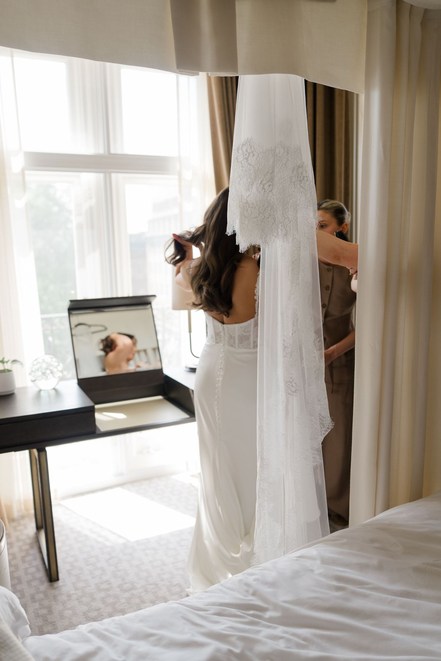 Bride in a white lace gown with cathedral veil stands before a mirror in a bright hotel room on her wedding day.
