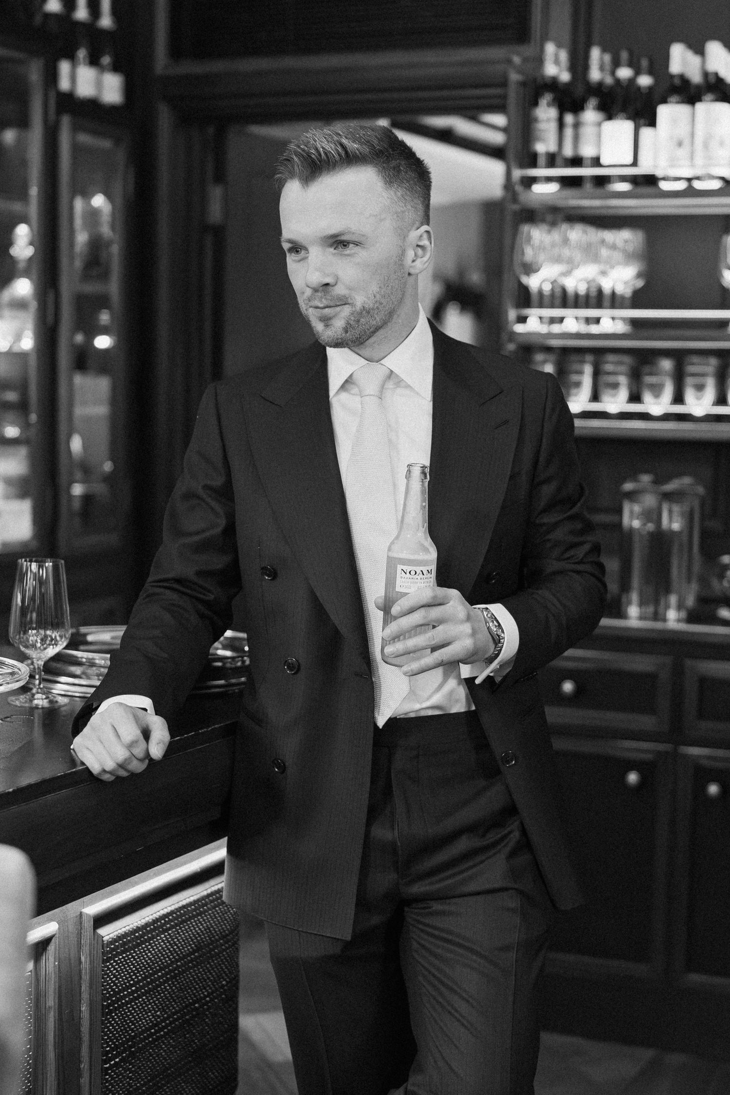Stylish man in black suit holding a bottle at an upscale bar in a black and white photo.