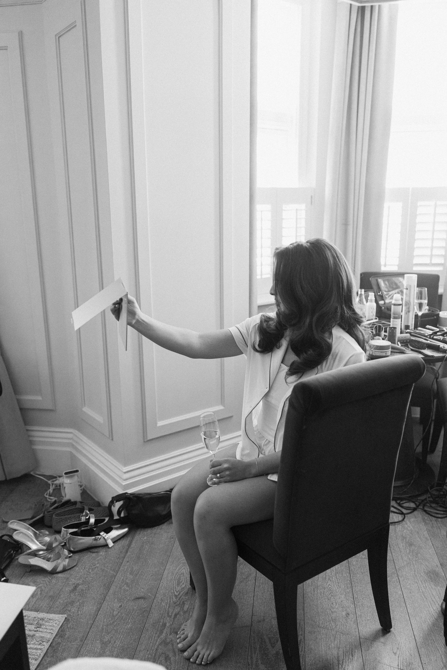 Black and white photo of a bride in pajamas reading a card while sitting in a chair during wedding morning preparations.