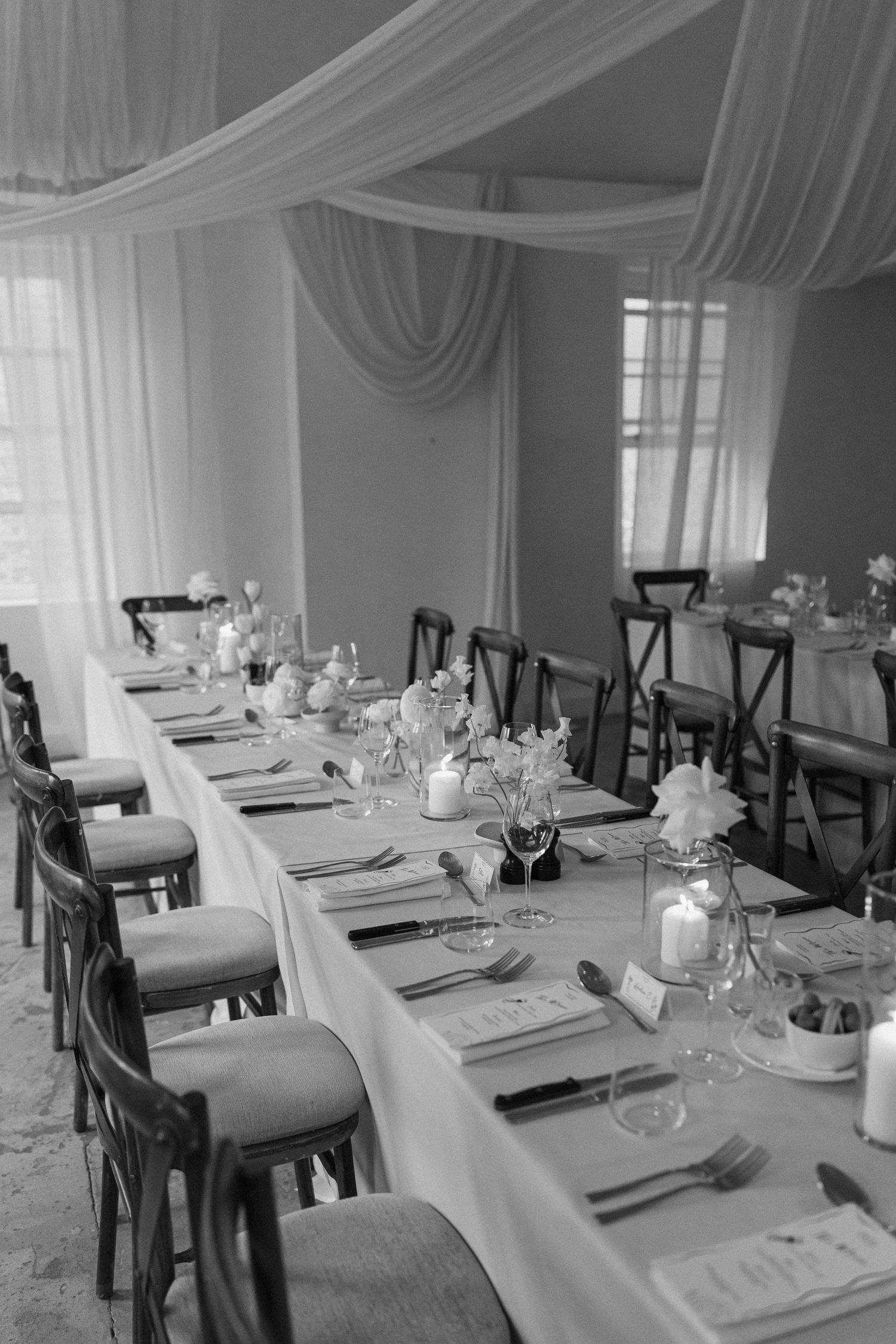 Black and white photo of a long elegantly set wedding reception table with draped ceiling decor.