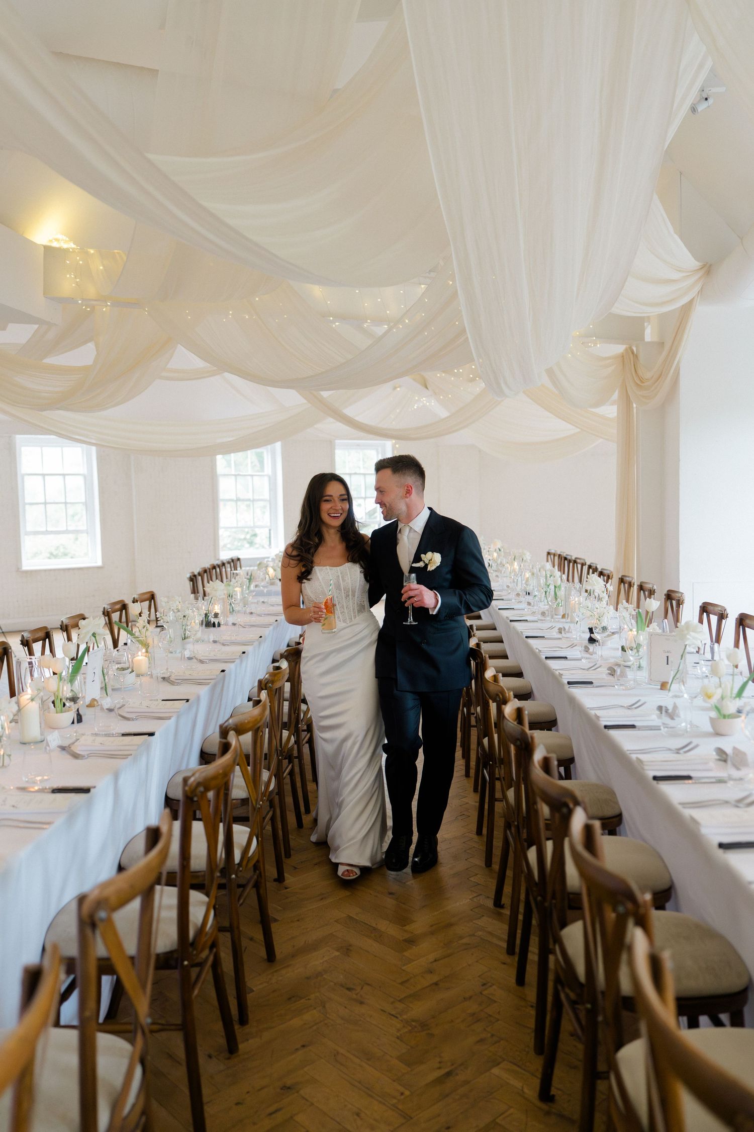 Bride and groom walk between reception tables adorned with white draping and floral centerpieces.