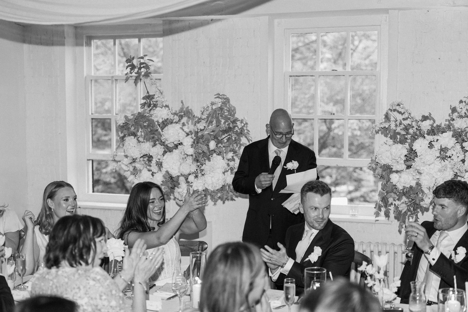 Bald man delivers heartfelt speech at wedding reception dinner table surrounded by guests and flowers.