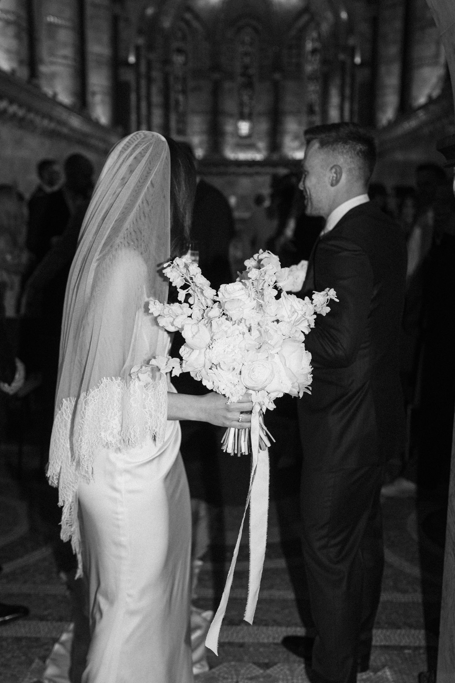 Black and white photo of bride in lace veil holding white floral bouquet with ribbons facing groom at altar.