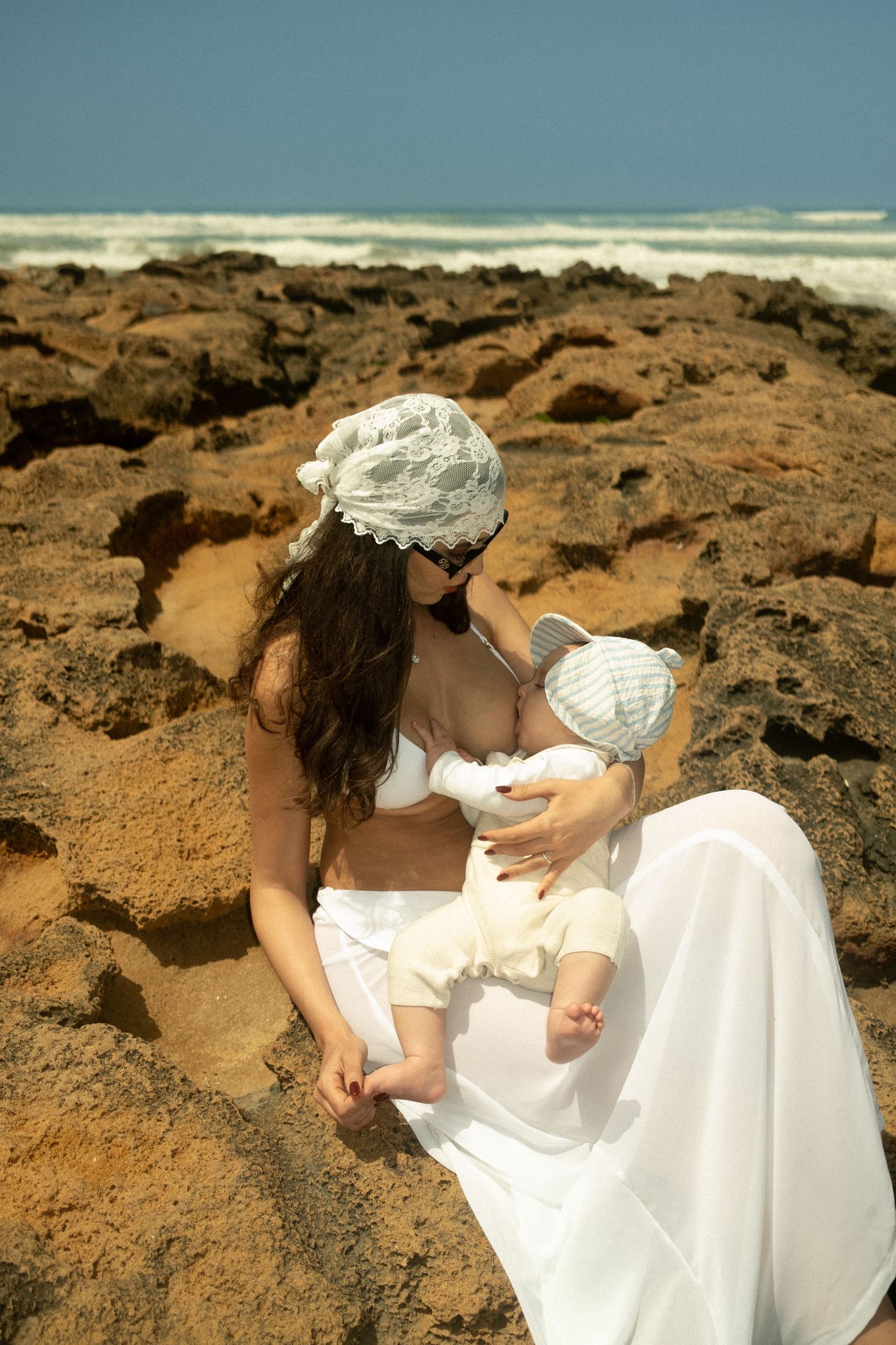 Mother in white dress and lace bonnet breastfeeding baby on rocky beach with ocean waves backdrop.