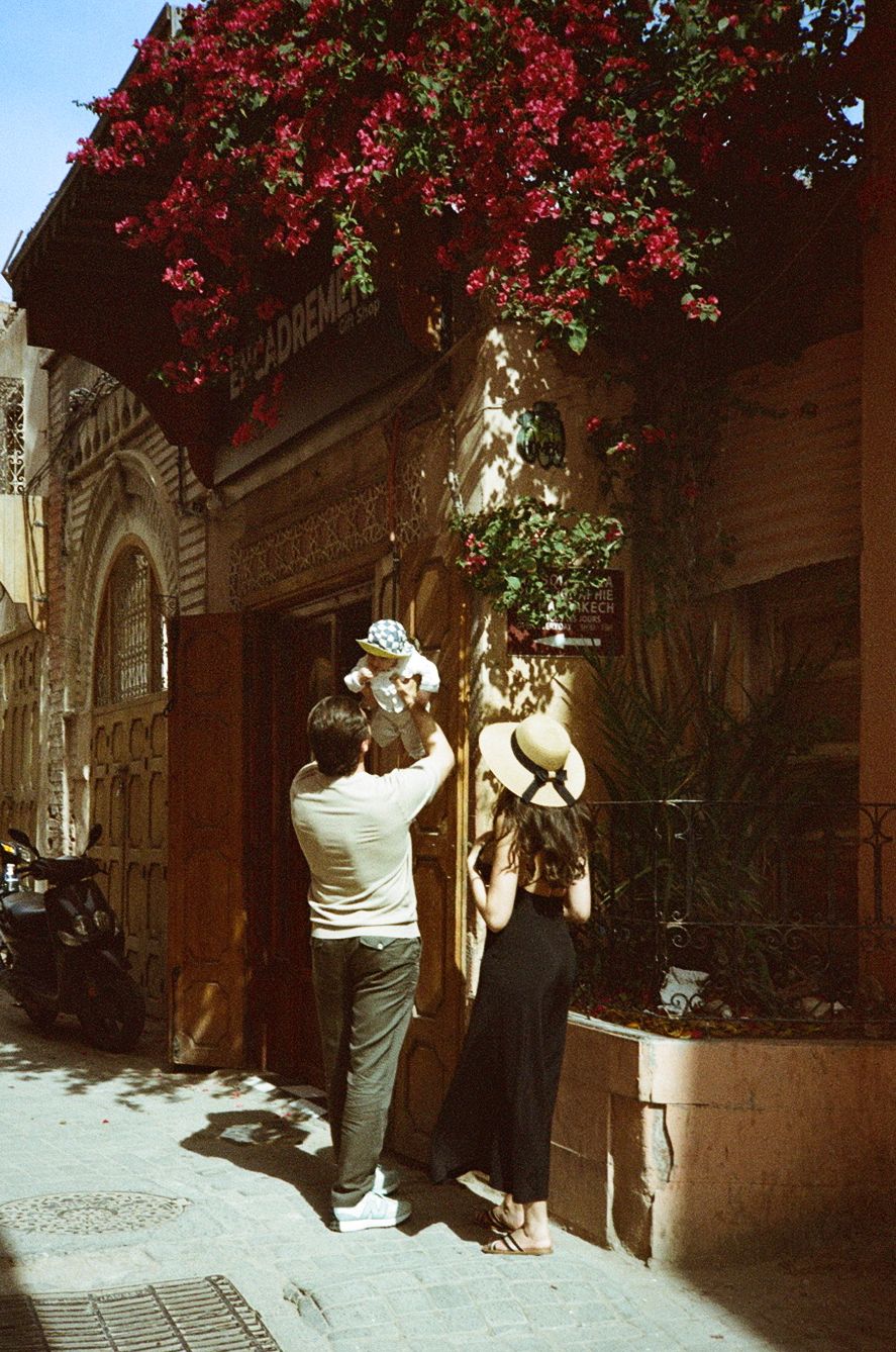 Couple photographing a doorway adorned with vibrant red bougainvillea flowers on a sunny street in a historic city.