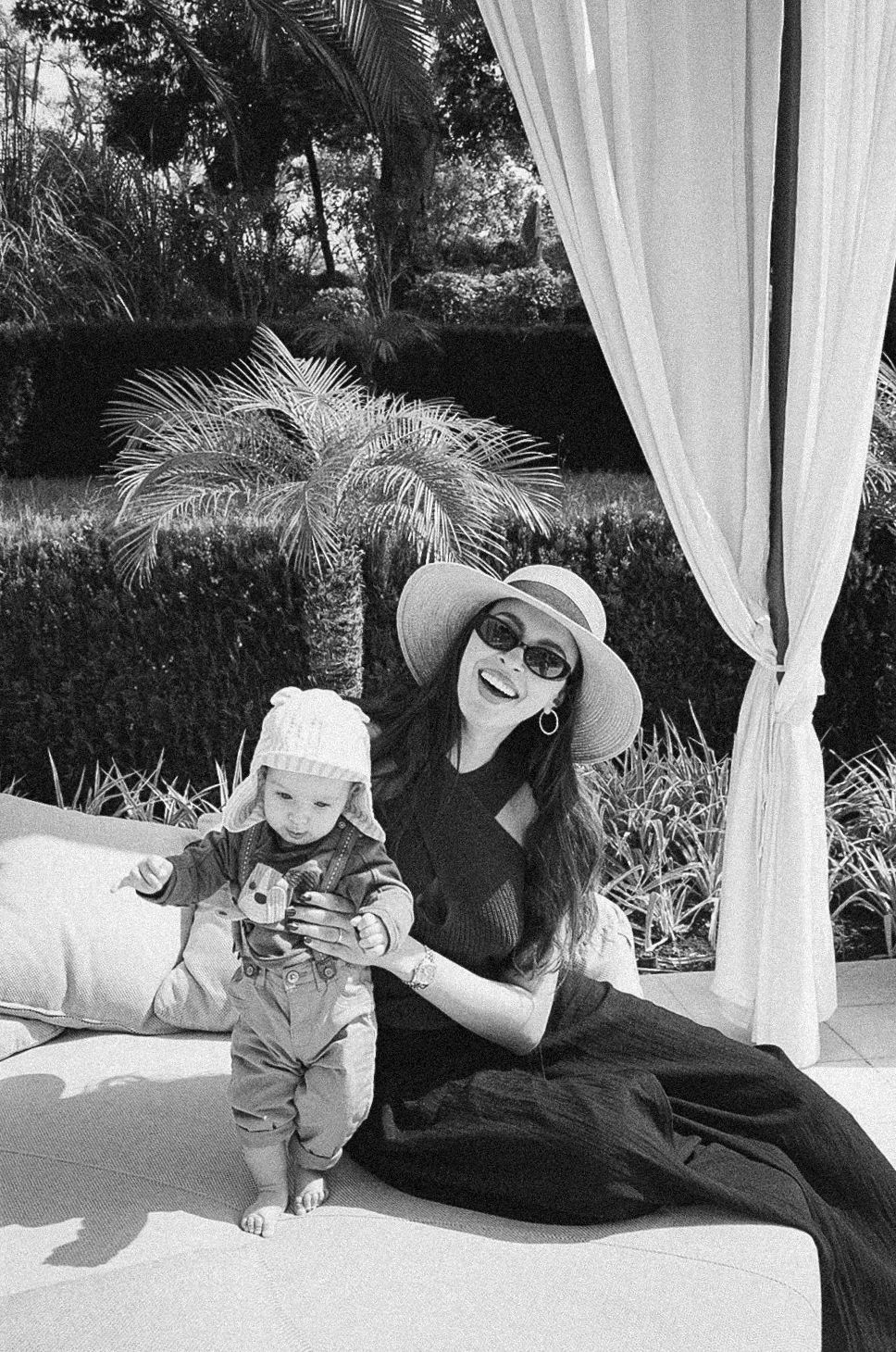 Black and white photo of a smiling woman in a sun hat and sunglasses sitting with a baby in a garden cabana.