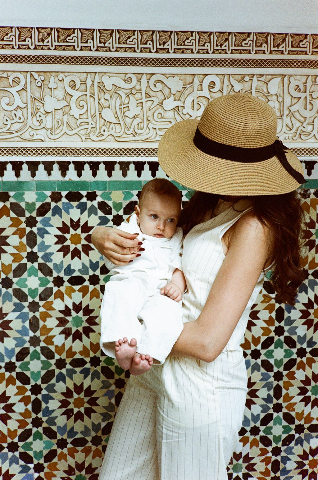 Woman in white outfit and sun hat holding a baby against colorful Moroccan mosaic tile wall.