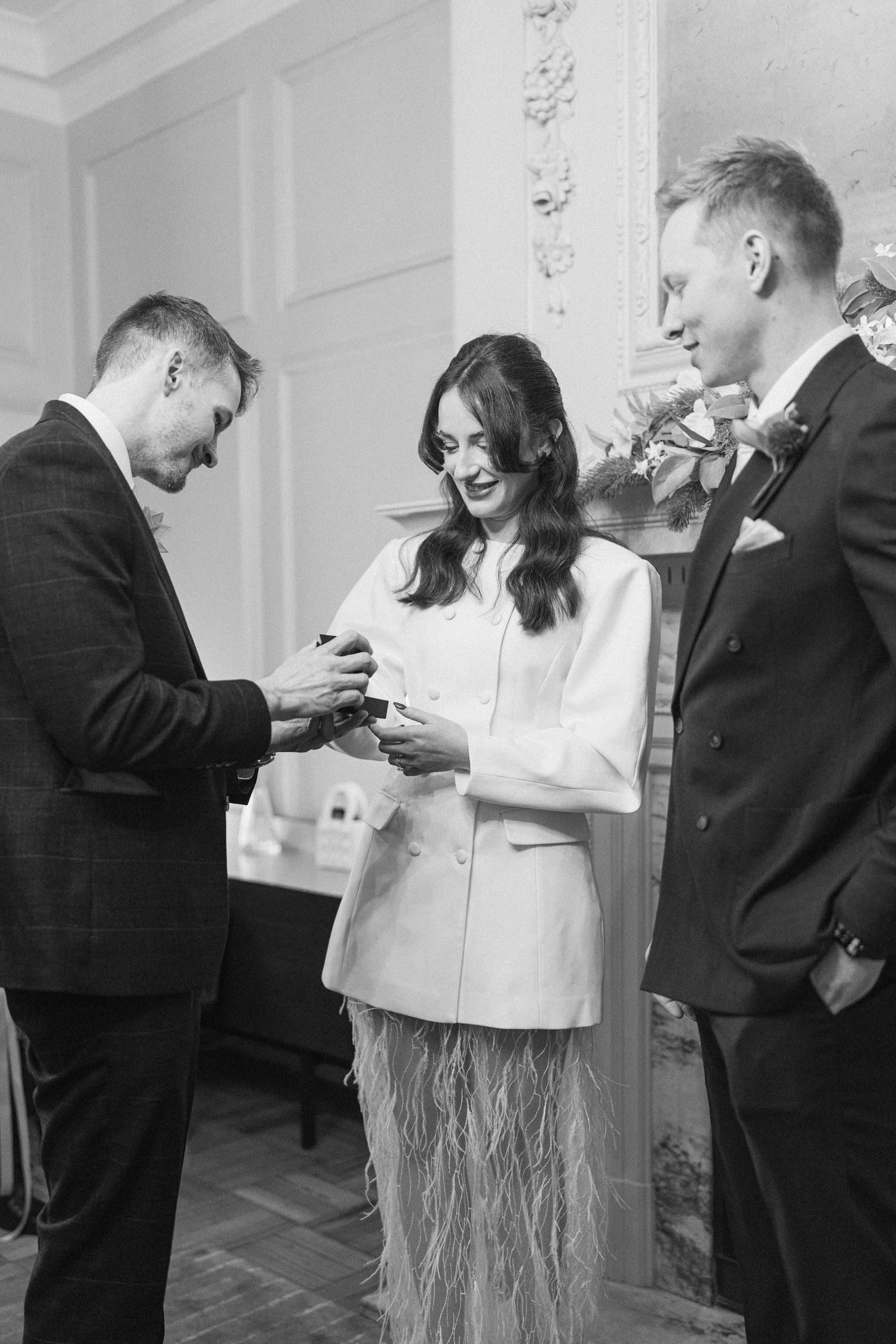 Black and white photo of a wedding officiant presenting rings to a bride and groom at a ceremony.