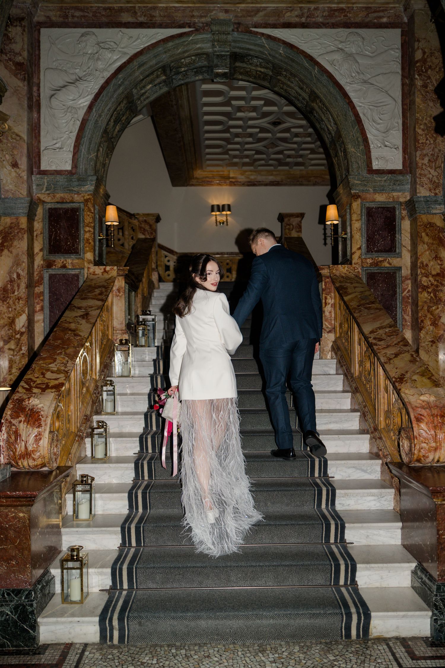 Bride in white feather skirt and groom in navy suit ascending grand marble staircase at elegant wedding venue.