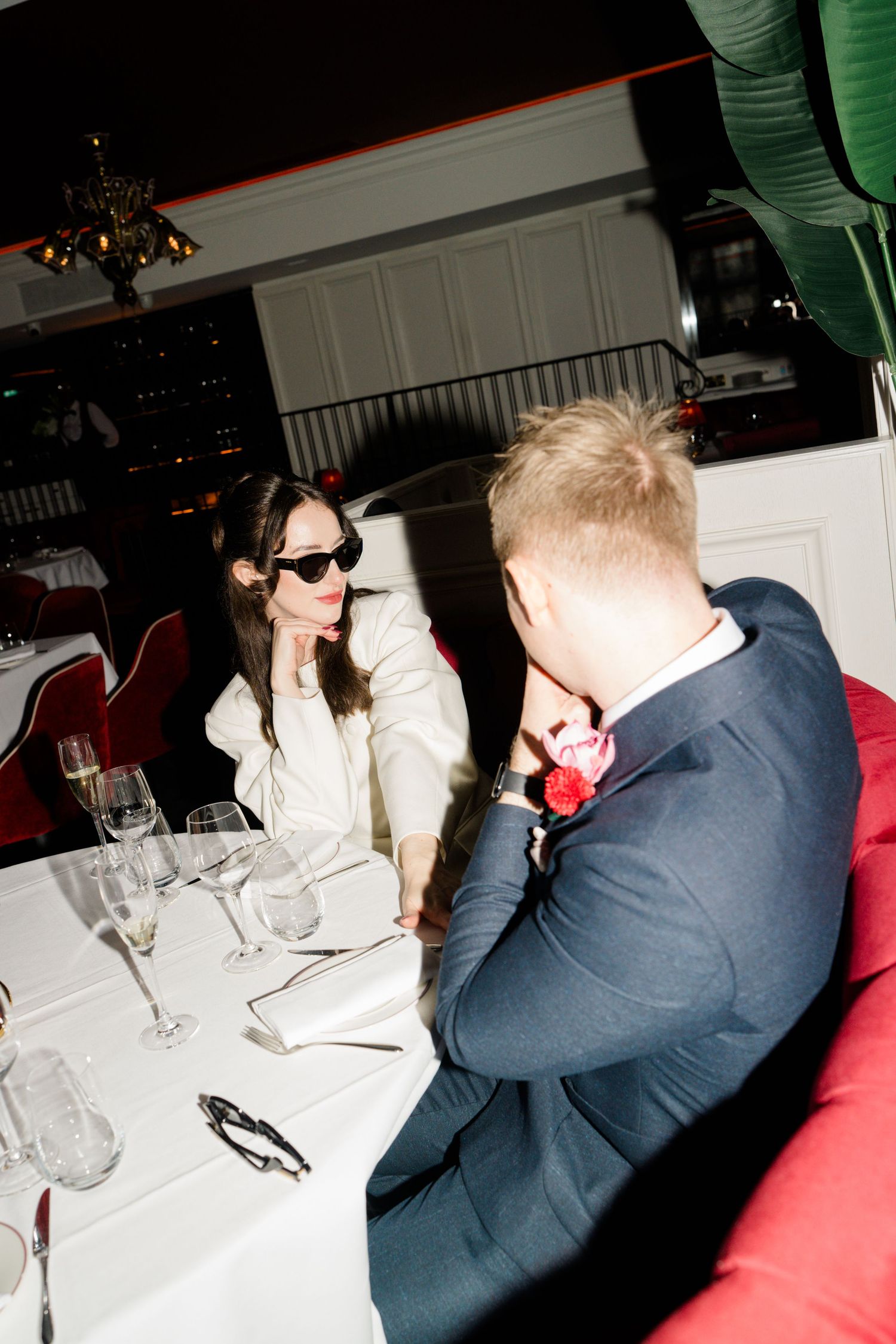 Bride in white suit wearing sunglasses sits with groom in navy suit at elegant restaurant table.