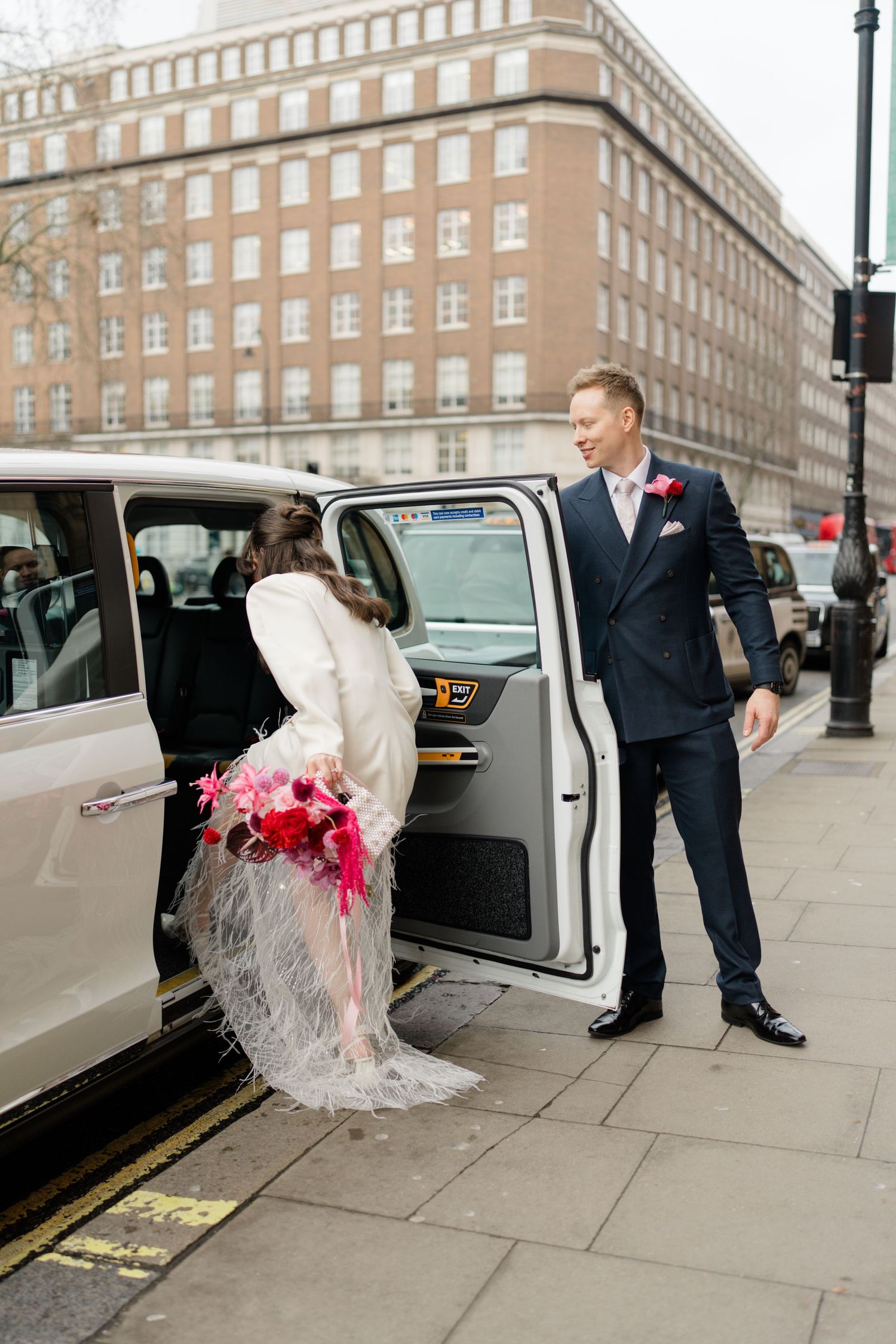 Groom in navy suit opening taxi door for bride in white feather skirt in London.