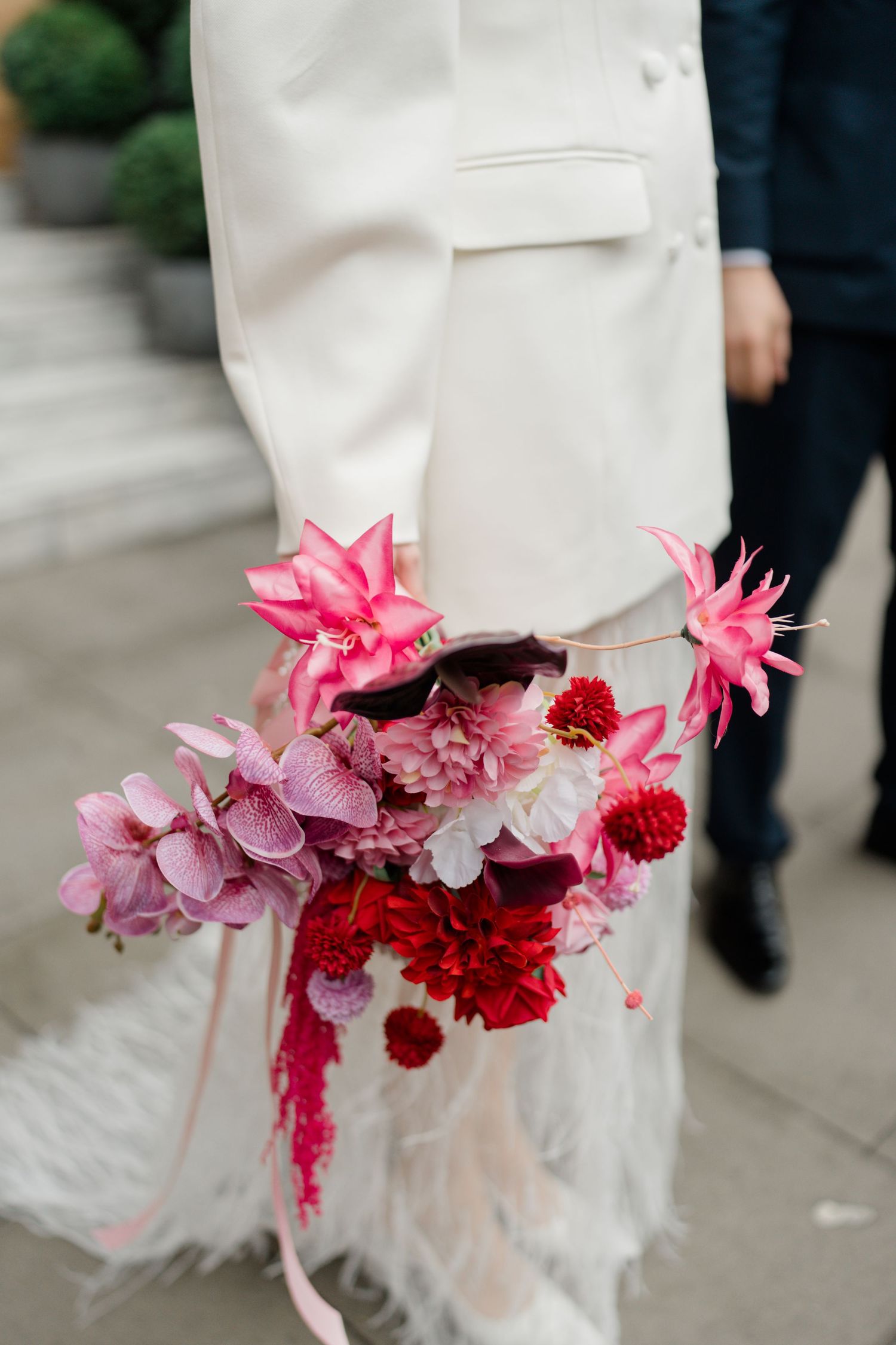 Bride in white feathered dress holds vibrant pink and red wedding bouquet with tropical flowers and ribbon streamers.