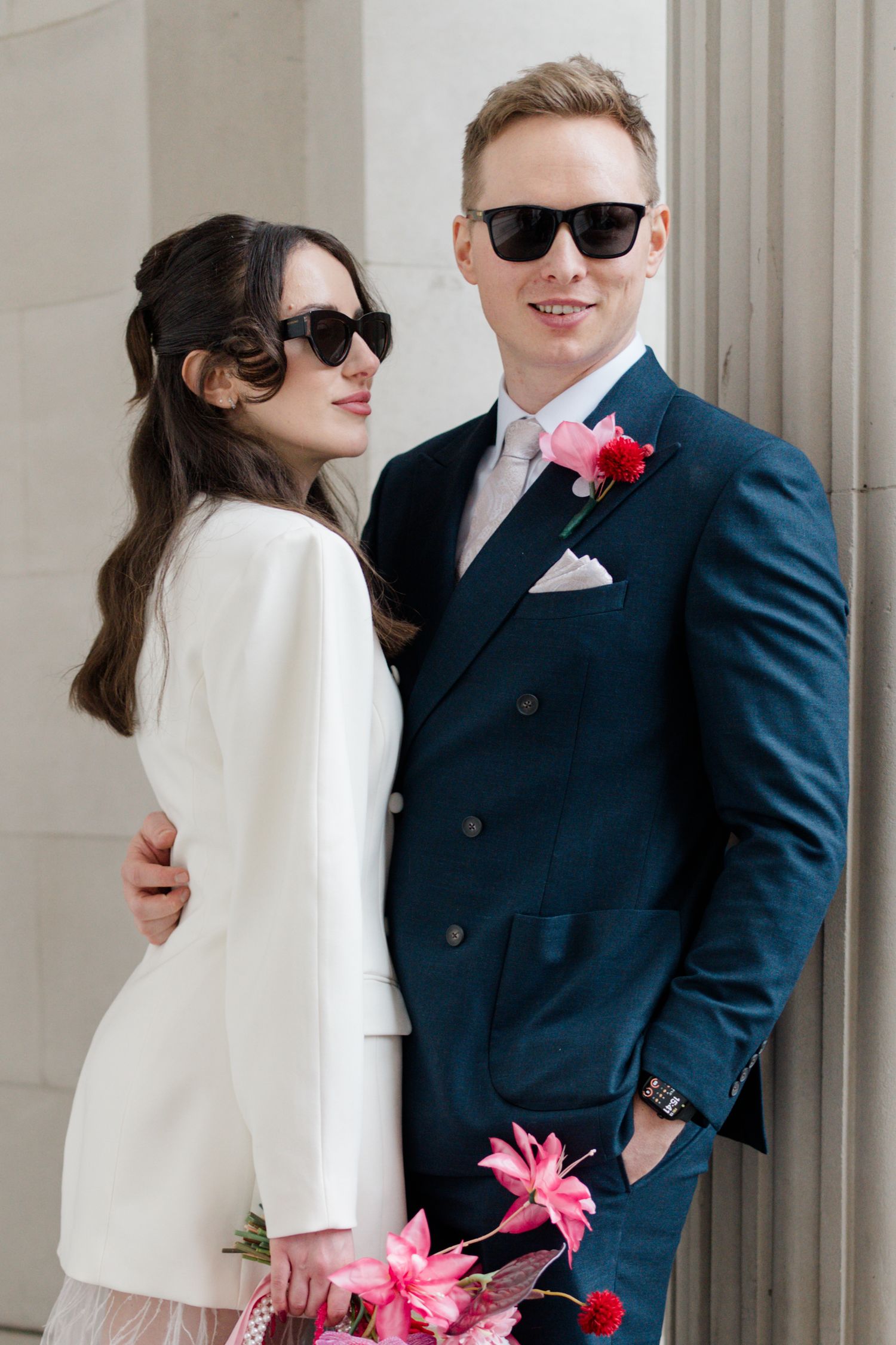 Stylish bride and groom wearing sunglasses with pink floral bouquet outdoors.