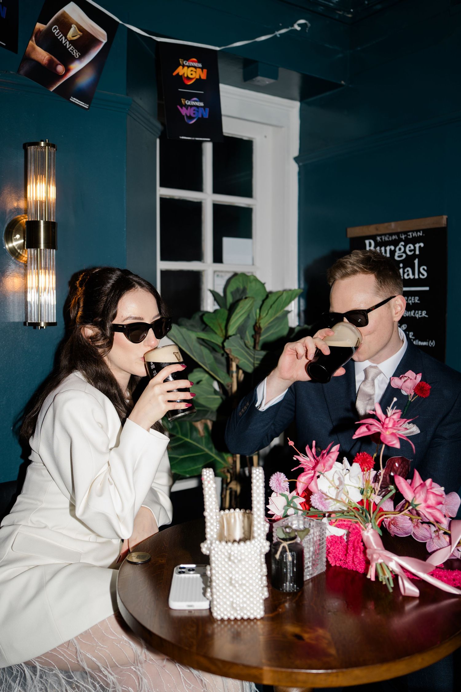 Bride and groom wearing sunglasses enjoy drinks at a pub table with pink floral centerpiece on wedding day.