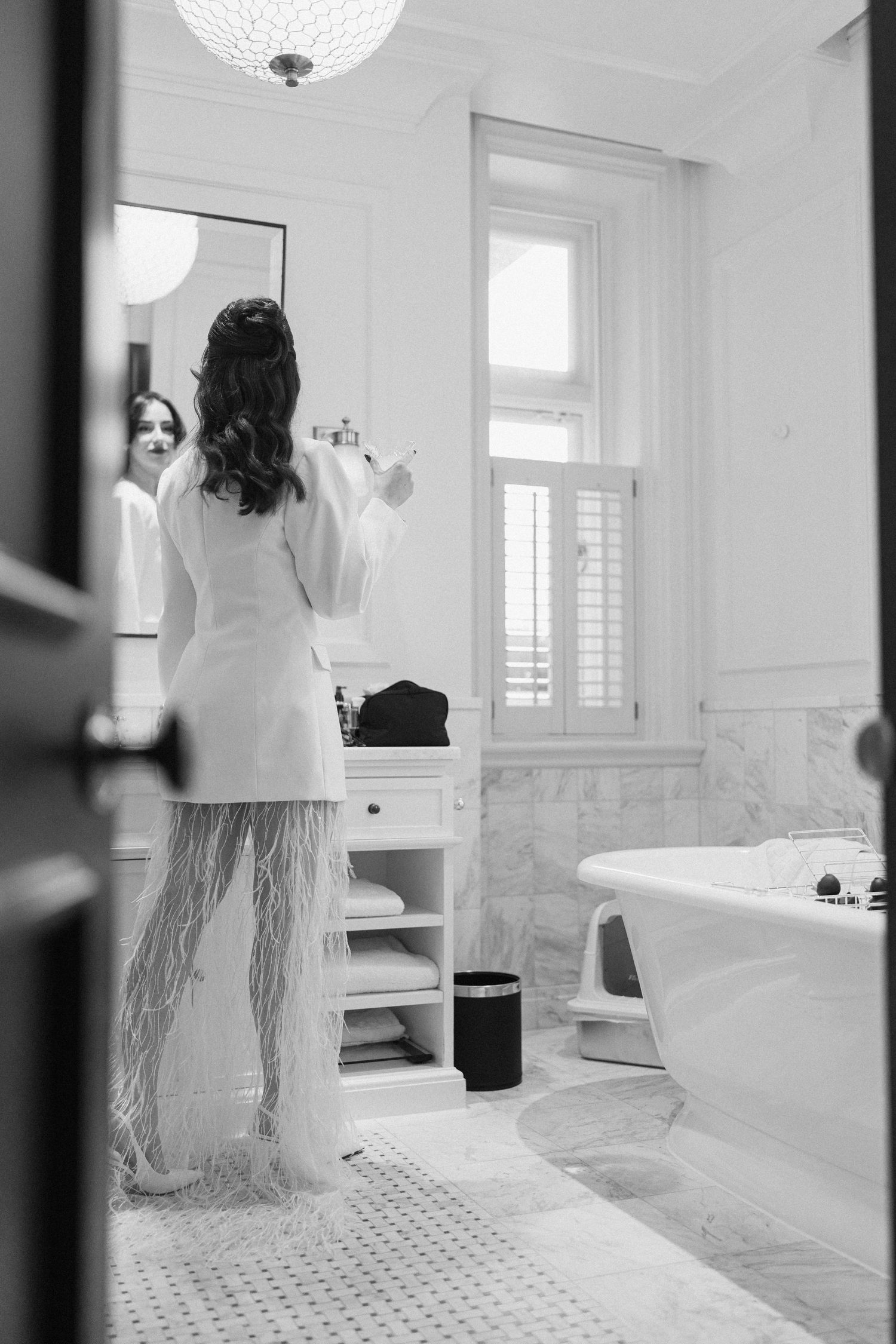 Bride in white blazer and feathered skirt stands before mirror in luxurious marble bathroom, black and white.