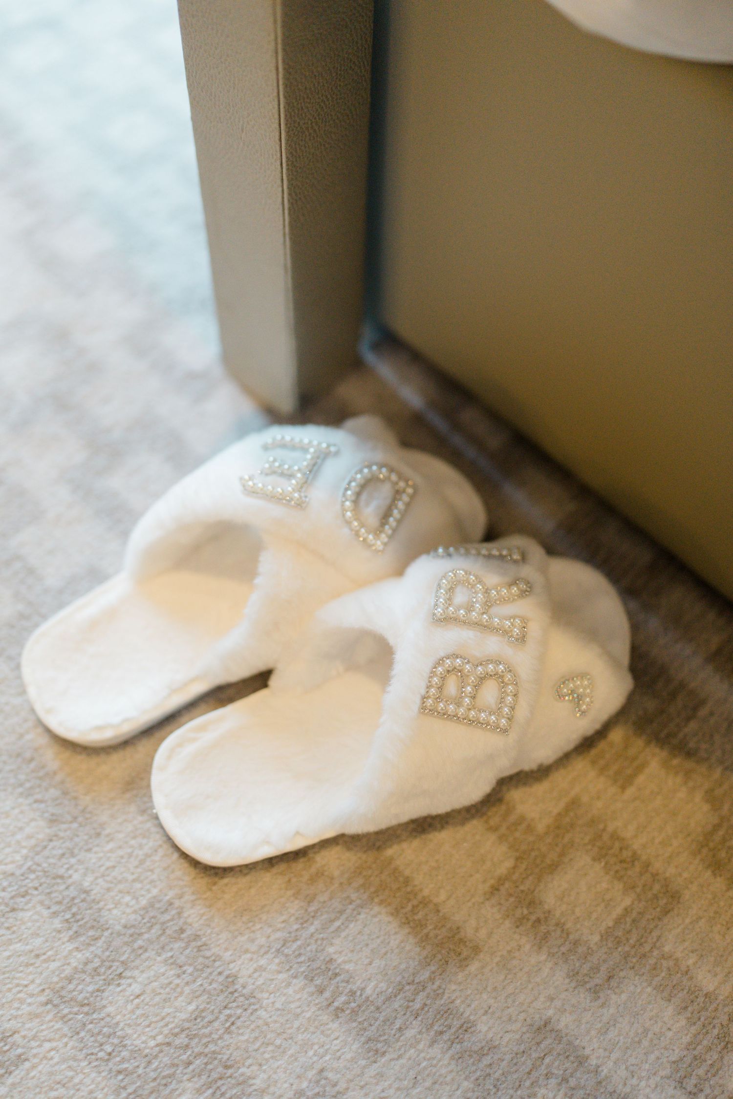 White fluffy bride slippers with pearl letter embellishments on hotel room floor.