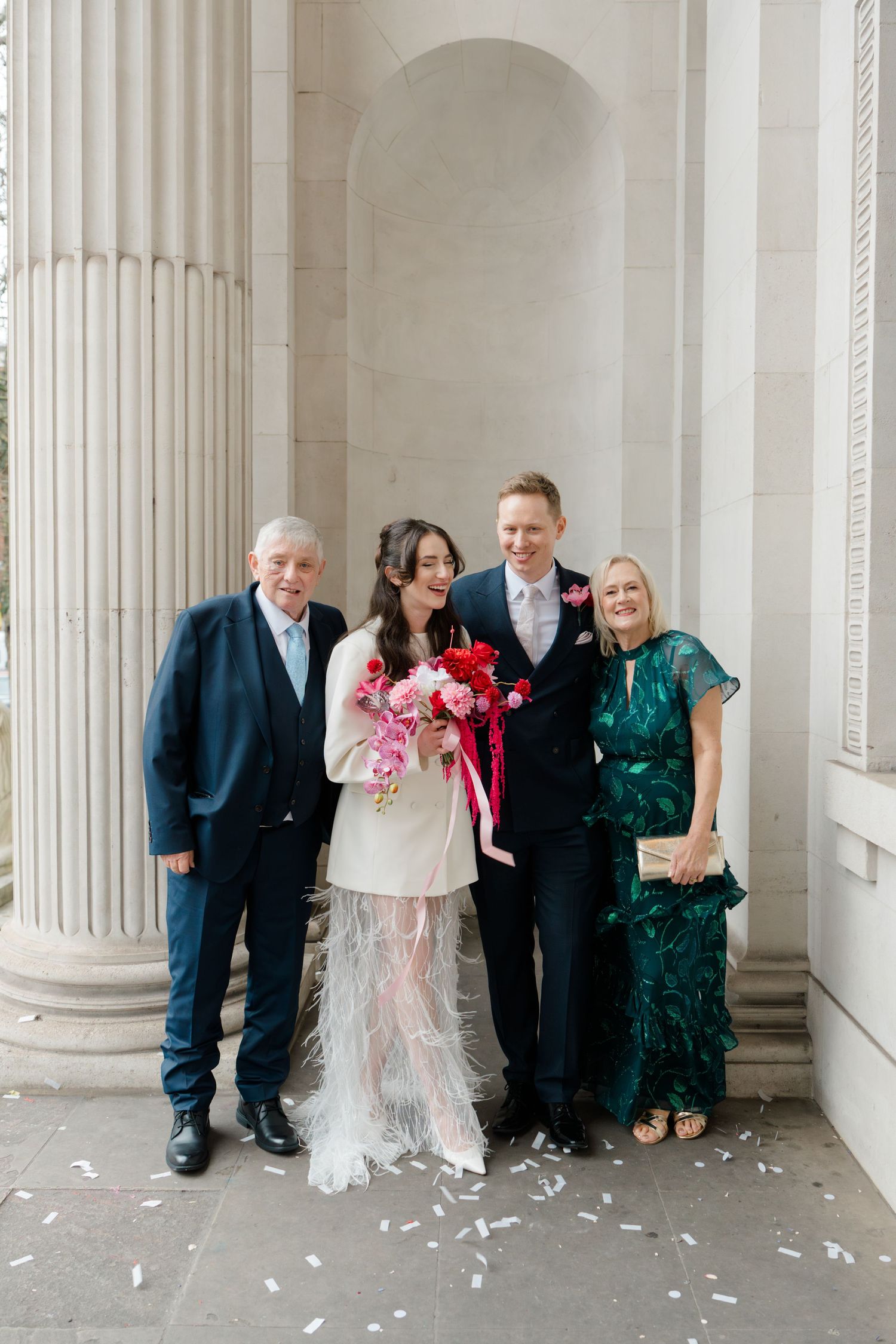 Wedding couple with parents posing between stone columns after ceremony with confetti.