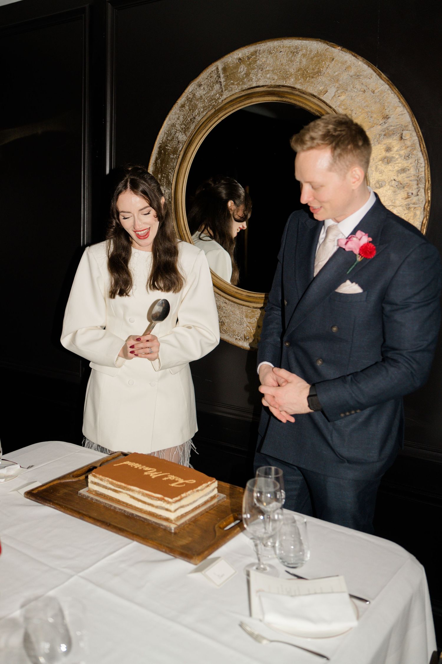 Bride in white coat and groom in navy suit prepare to cut Just Married chocolate wedding cake.
